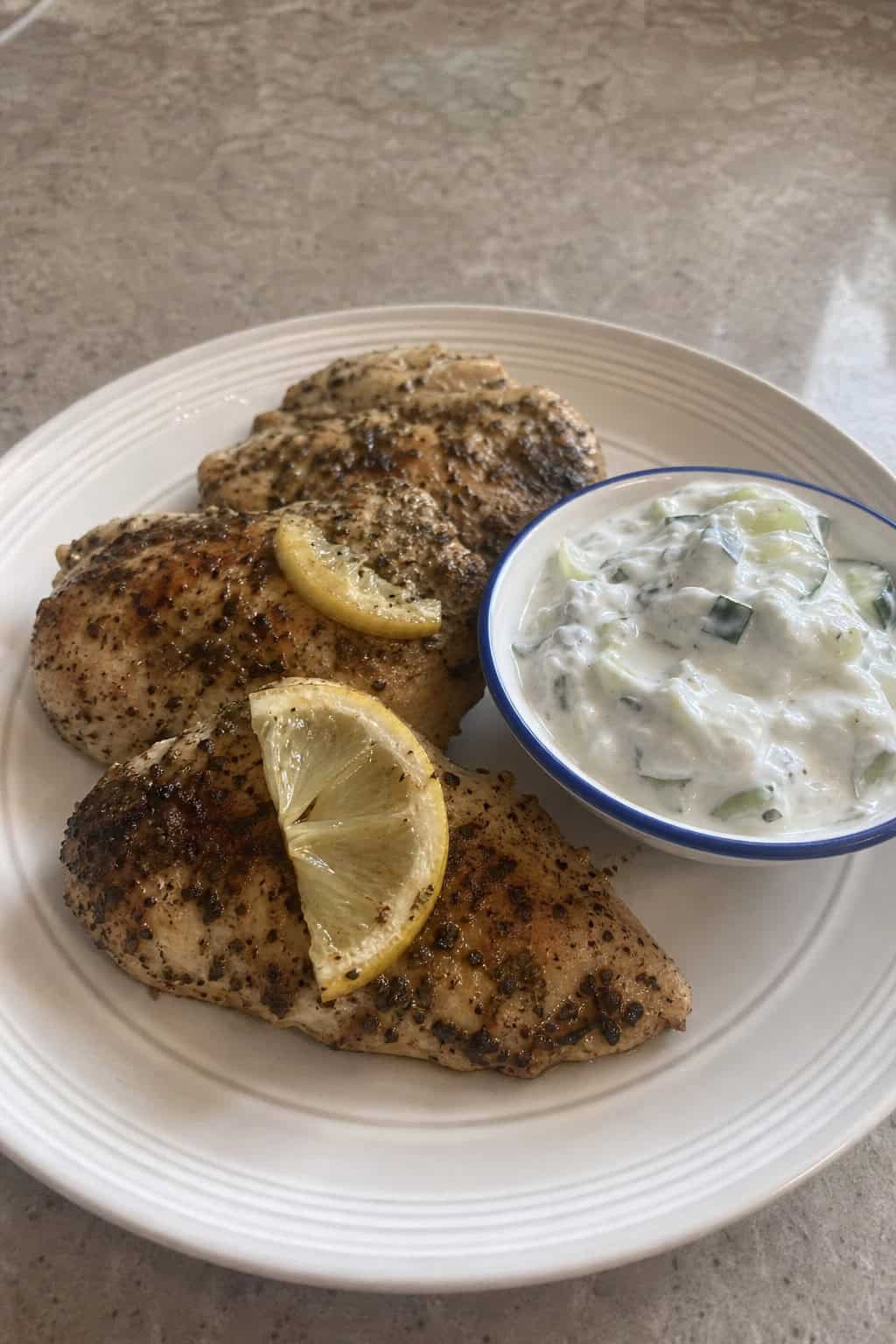 A plate of golden lemon-oregano chicken served with cucumber yogurt sauce and fresh herb garnishes on a wooden table.