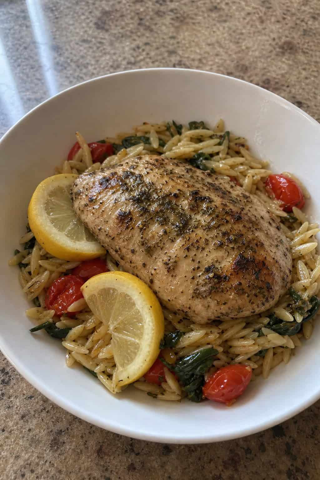 A bowl of lemon-oregano chicken served on orzo pasta with fresh herbs and lemon slices on a wooden table.