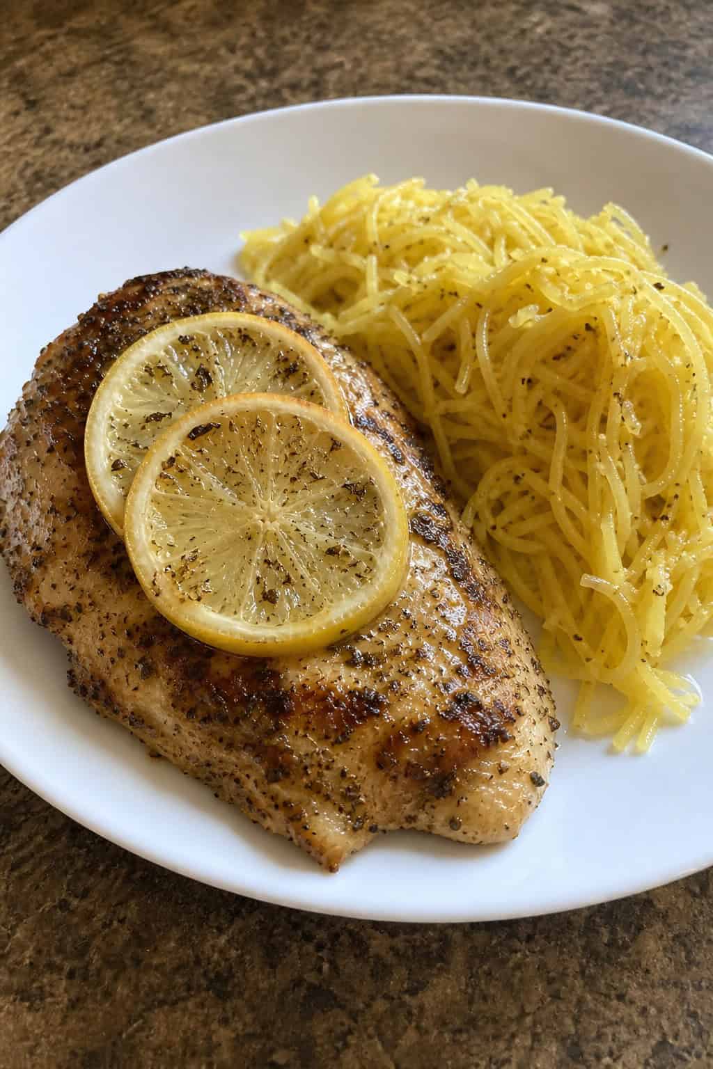 A plate of golden-brown lemon-oregano chicken served with roasted spaghetti squash on a wooden table with fresh lemons and herbs in the background.