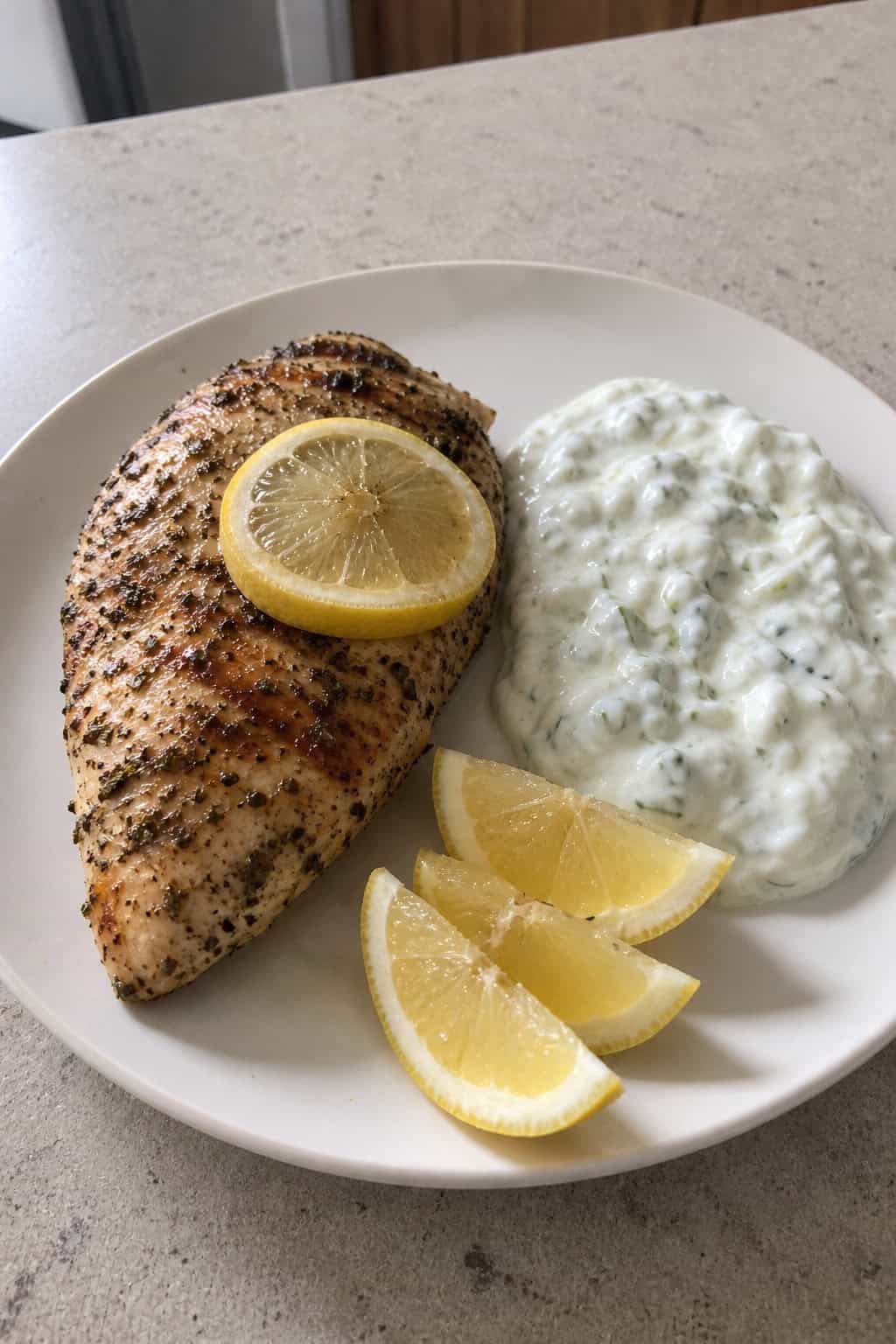 A plate of golden lemon-oregano chicken breast served with creamy tzatziki sauce and garnished with lemon wedges and oregano on a wooden table.