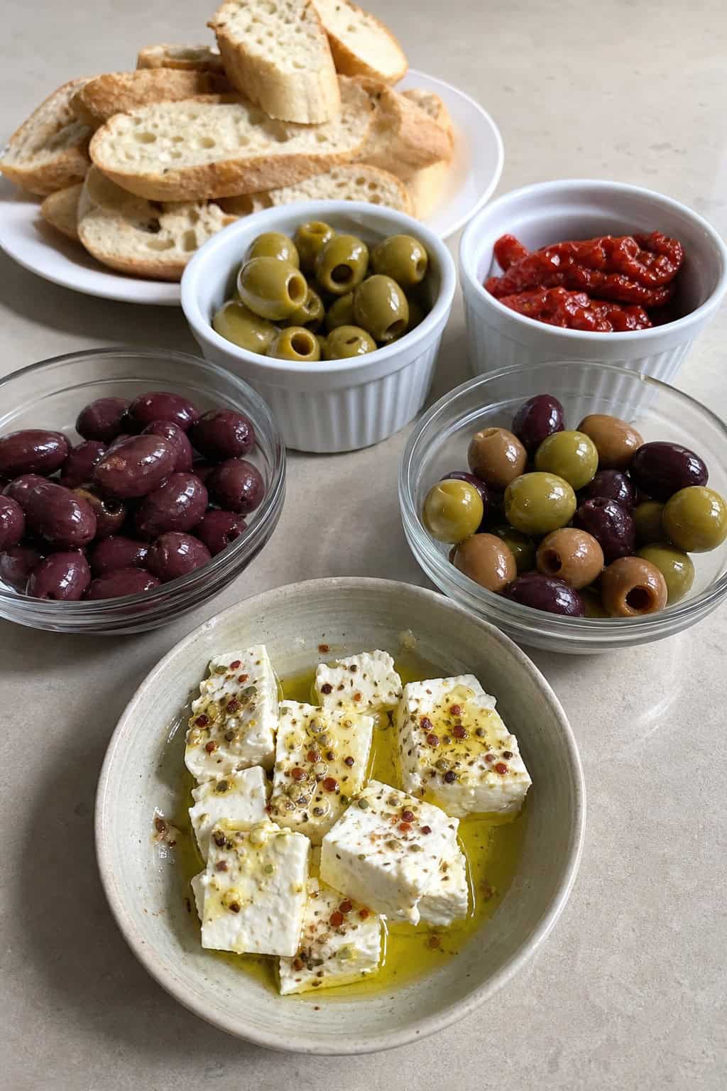 A Greek mezze platter with marinated feta cheese, olives, bread slices, and fresh herbs arranged on a wooden board.