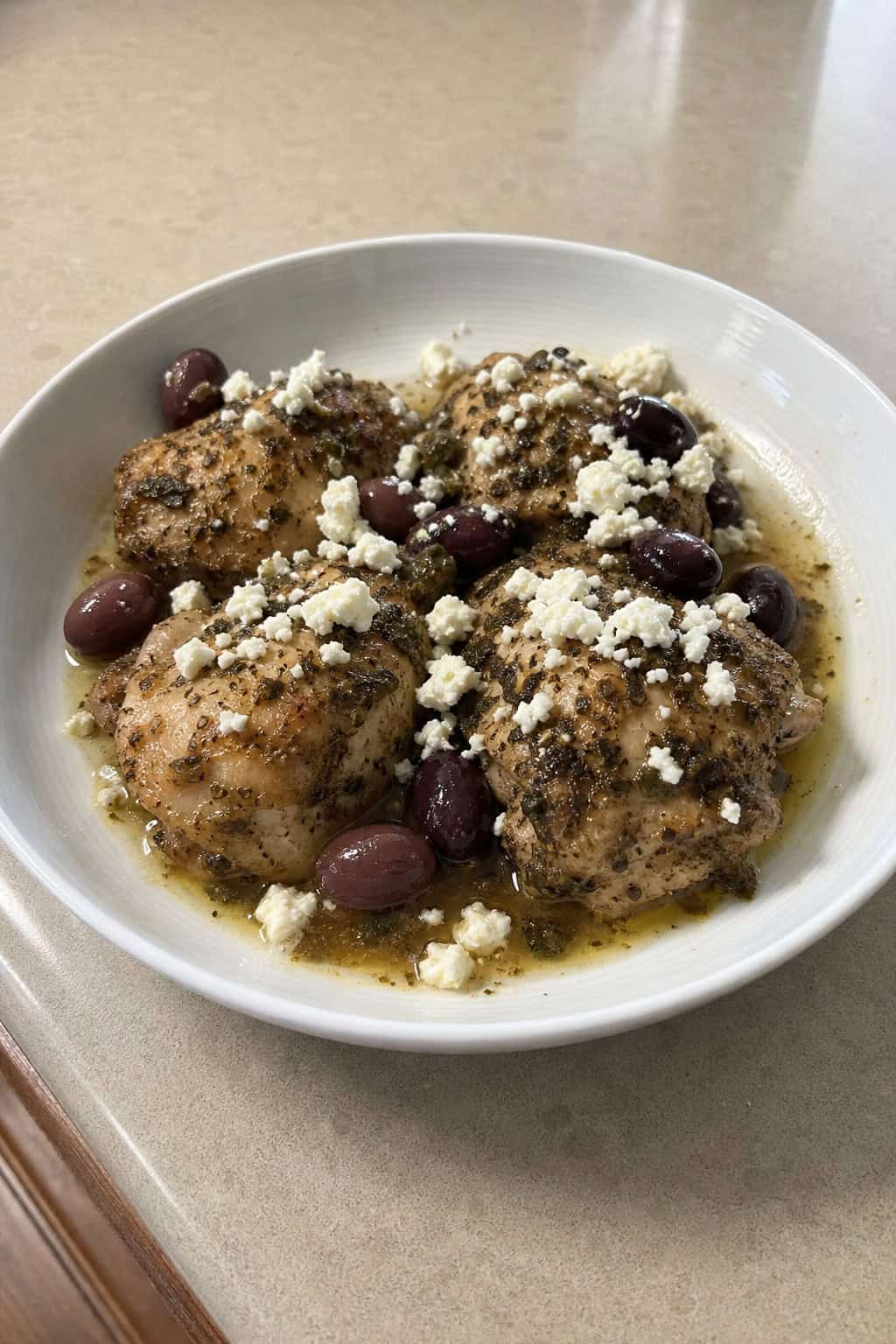 A bowl of cooked chicken with feta cheese and olives, garnished with herbs and lemon wedges on a kitchen table.