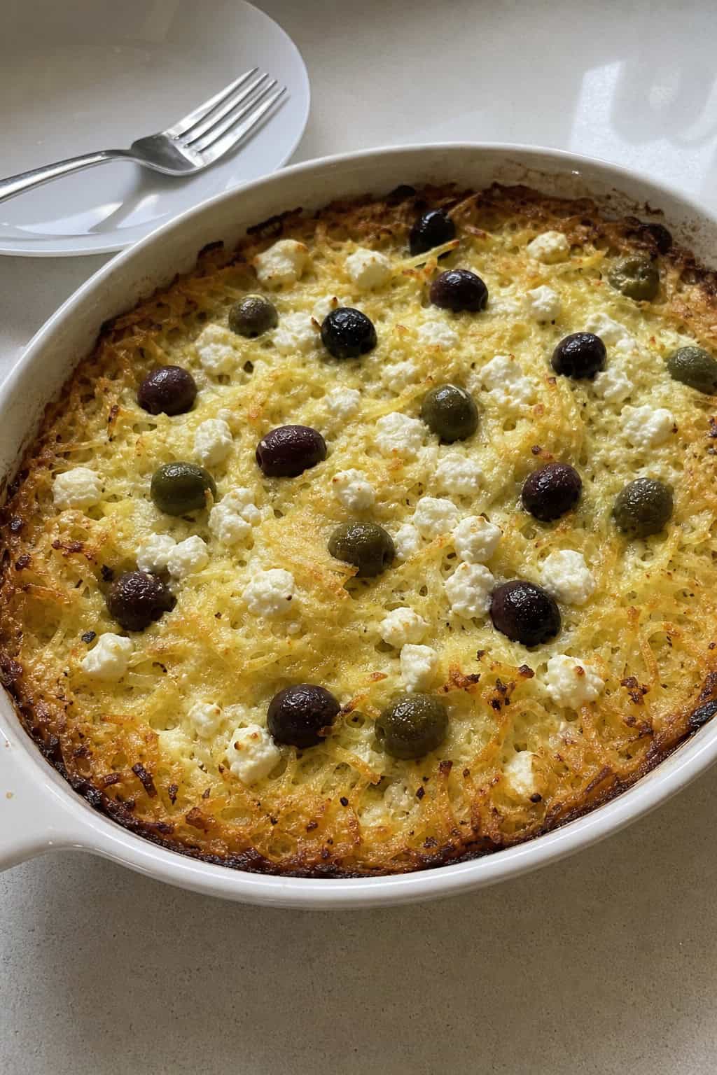 A baked casserole with golden hashbrowns, feta cheese, olives, and herbs in a ceramic dish on a wooden table.