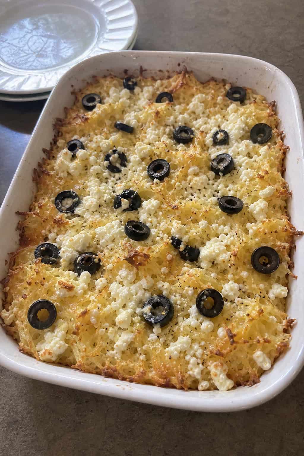 A baked hashbrown casserole with visible crumbles of white feta and sliced black olives on top, in a ceramic dish on a wood table with a small dish of extra feta nearby.