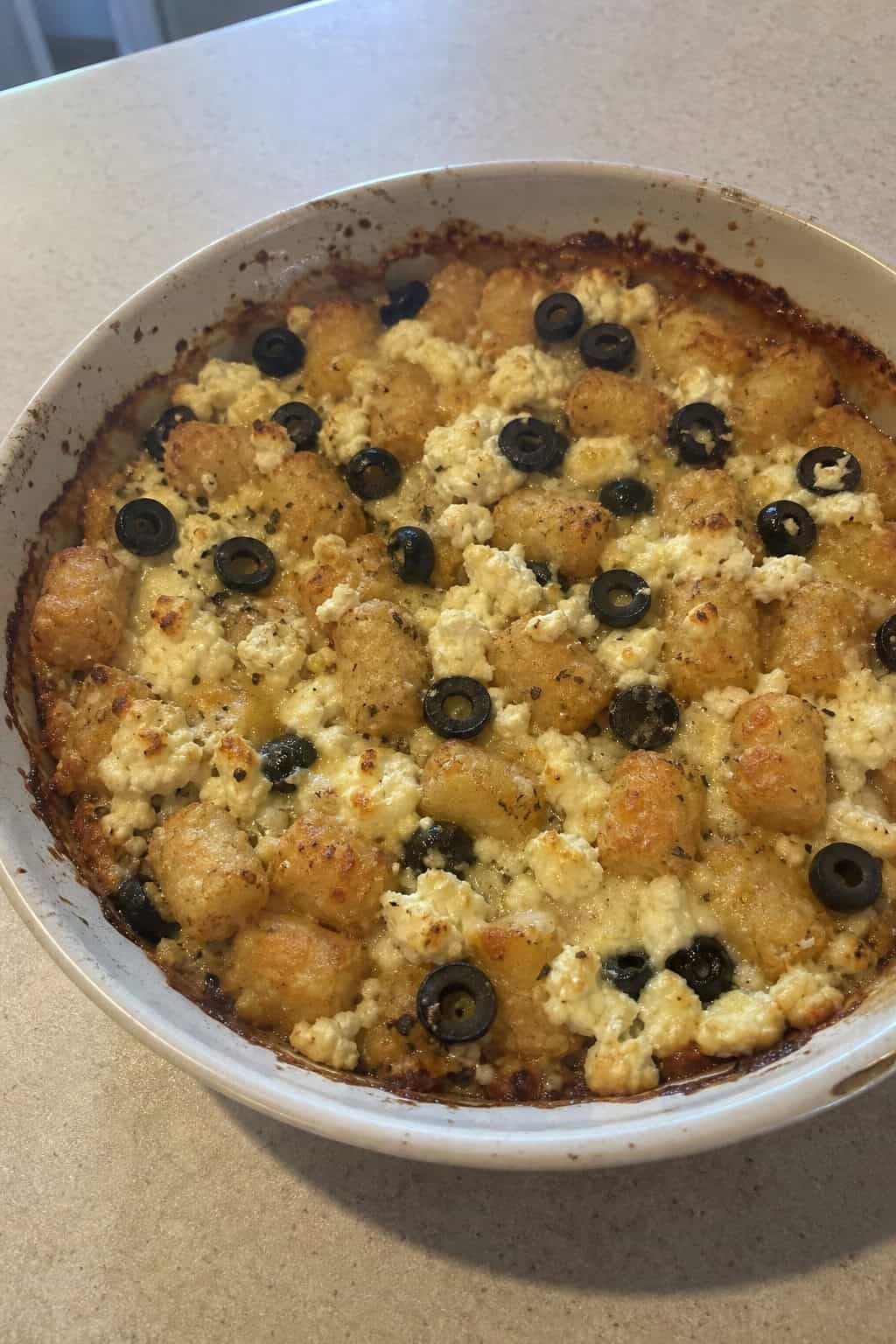 A golden brown tater tot casserole topped with crumbled white feta and dark Kalamata olives on a wooden table, with a bowl of olives and a fresh green salad alongside it.