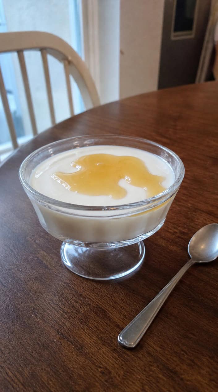 A glass dish of Greek yogurt and honey panna cotta topped with honey and garnished with mint leaves on a wooden table in warm sunlight.