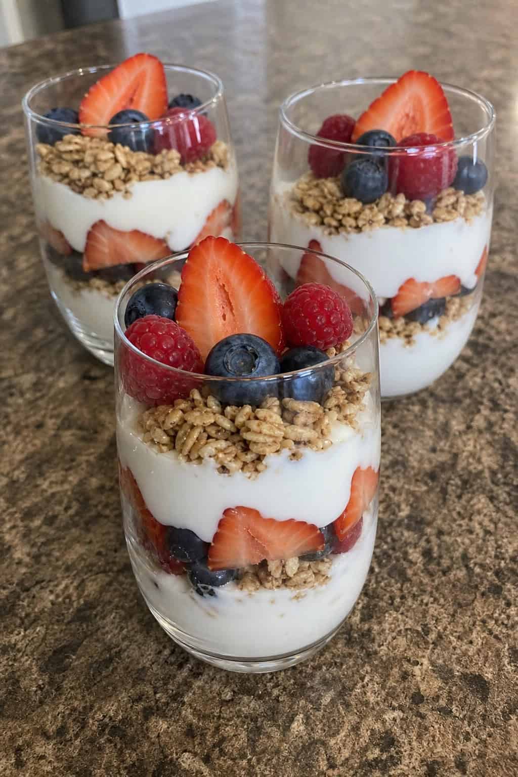 Three glass cups filled with layers of Greek yogurt, mixed berries, and granola on a wooden table with fresh berries around.