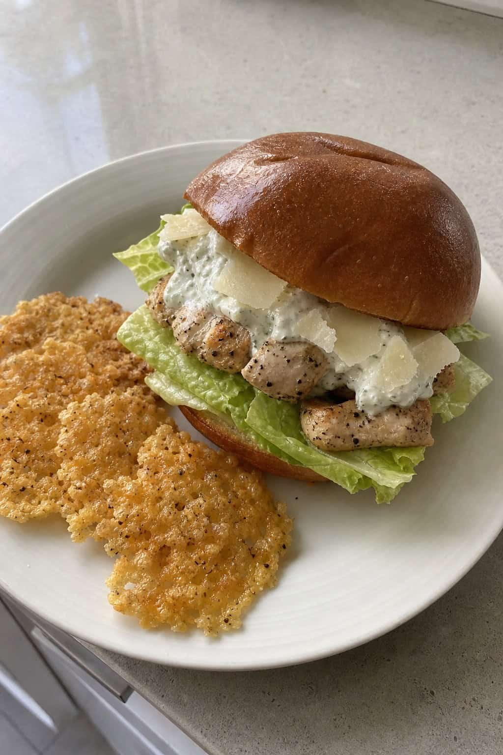 A grilled chicken sandwich with romaine lettuce and golden Parmesan crisps on a toasted bun, set on a wooden board