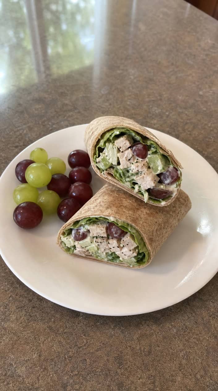 A halved chicken salad wrap with Greek yogurt filling on a wooden board surrounded by lettuce and grapes.