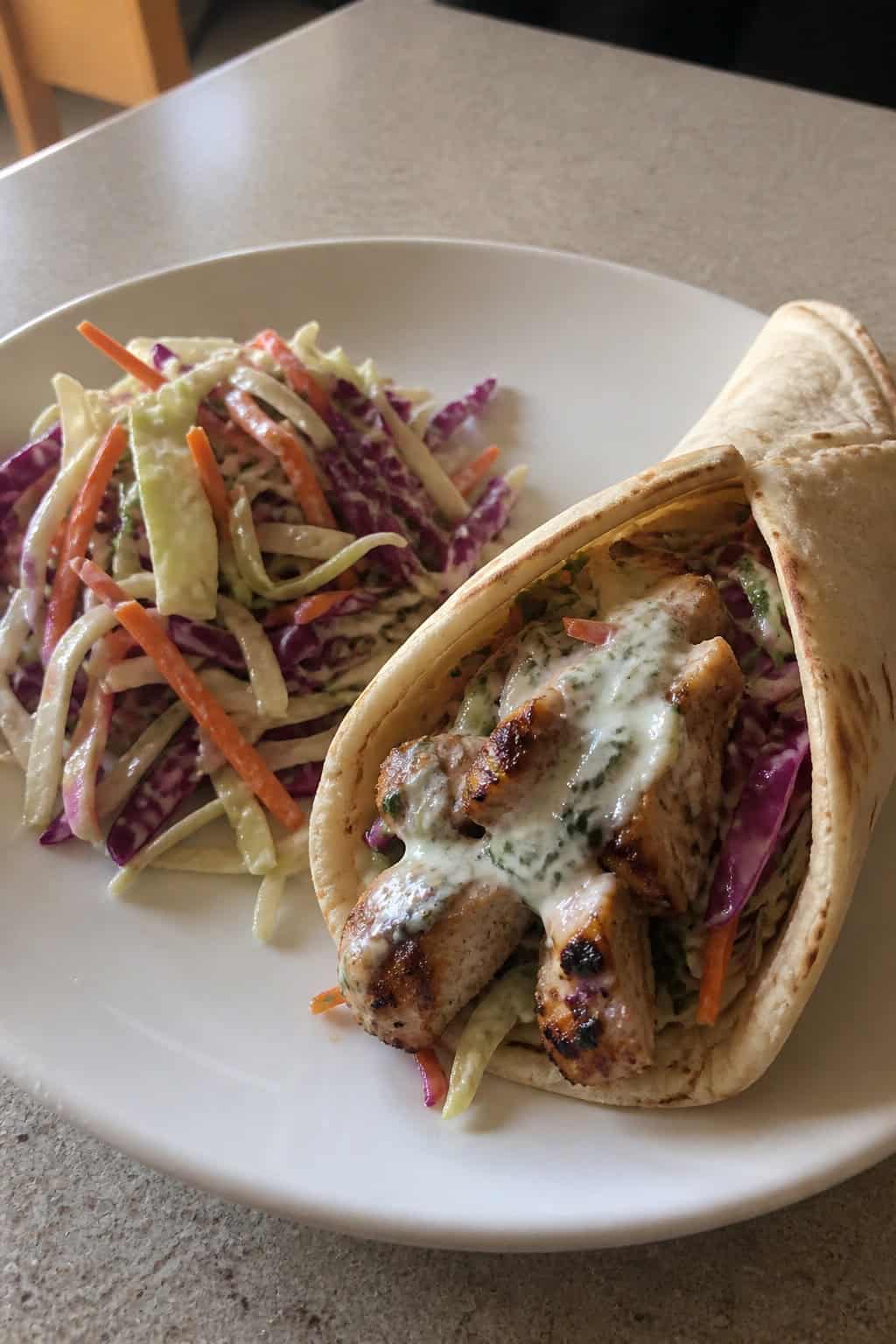 Close-up of a Greek yogurt marinated chicken gyro with cabbage slaw on a wooden board, accompanied by a small bowl of slaw and a lemon wedge.