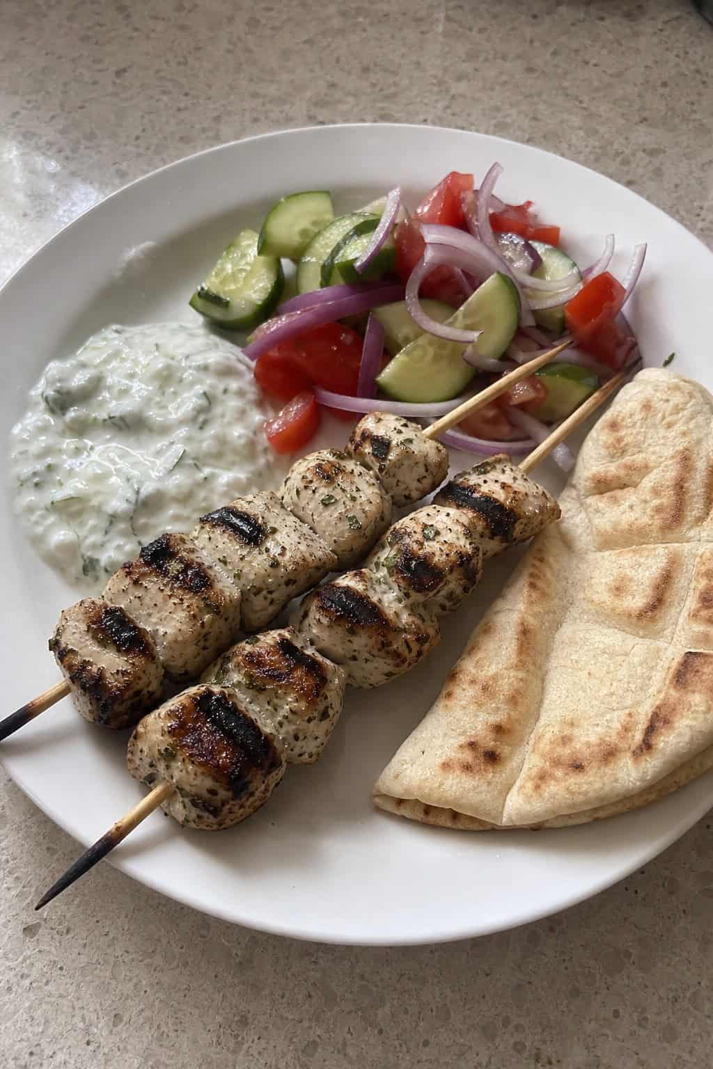 Grilled chicken souvlaki skewers with pita bread, fresh vegetables, and a bowl of tzatziki sauce on a wooden board.