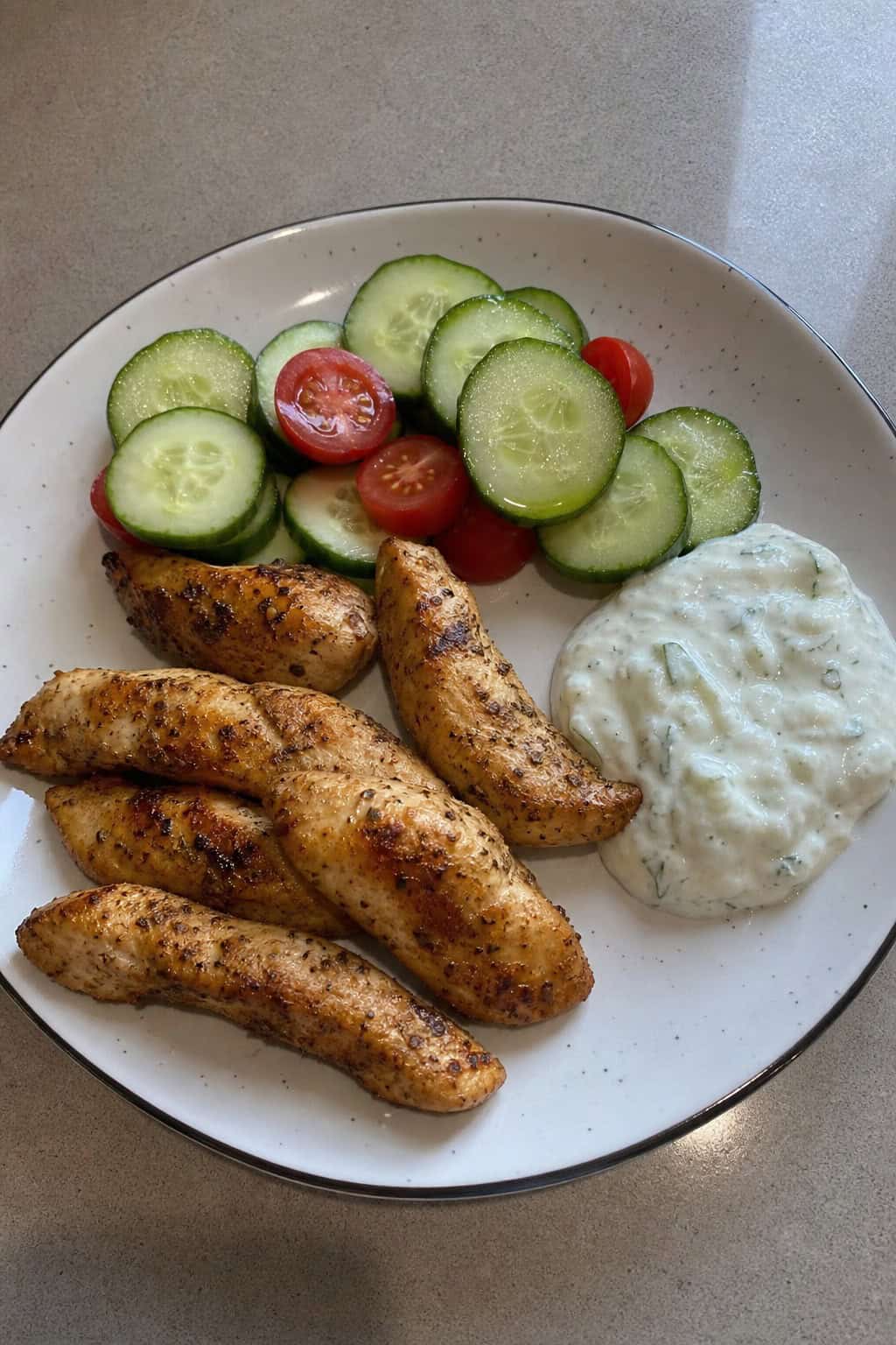 A plate of golden-brown chicken tenders with a bowl of creamy tzatziki sauce and fresh vegetable garnishes on a wooden table.
