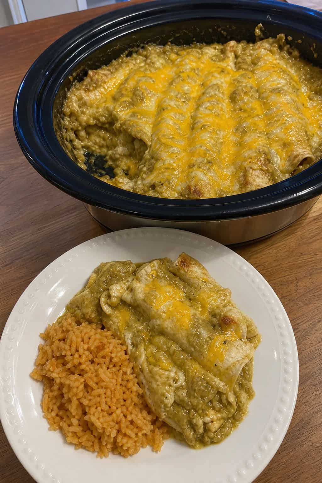 A crockpot insert filled with layered green chile and cheese enchilada casserole, with visible melted cheddar on top and fresh green chiles and shredded cheese on the kitchen countertop beside it.