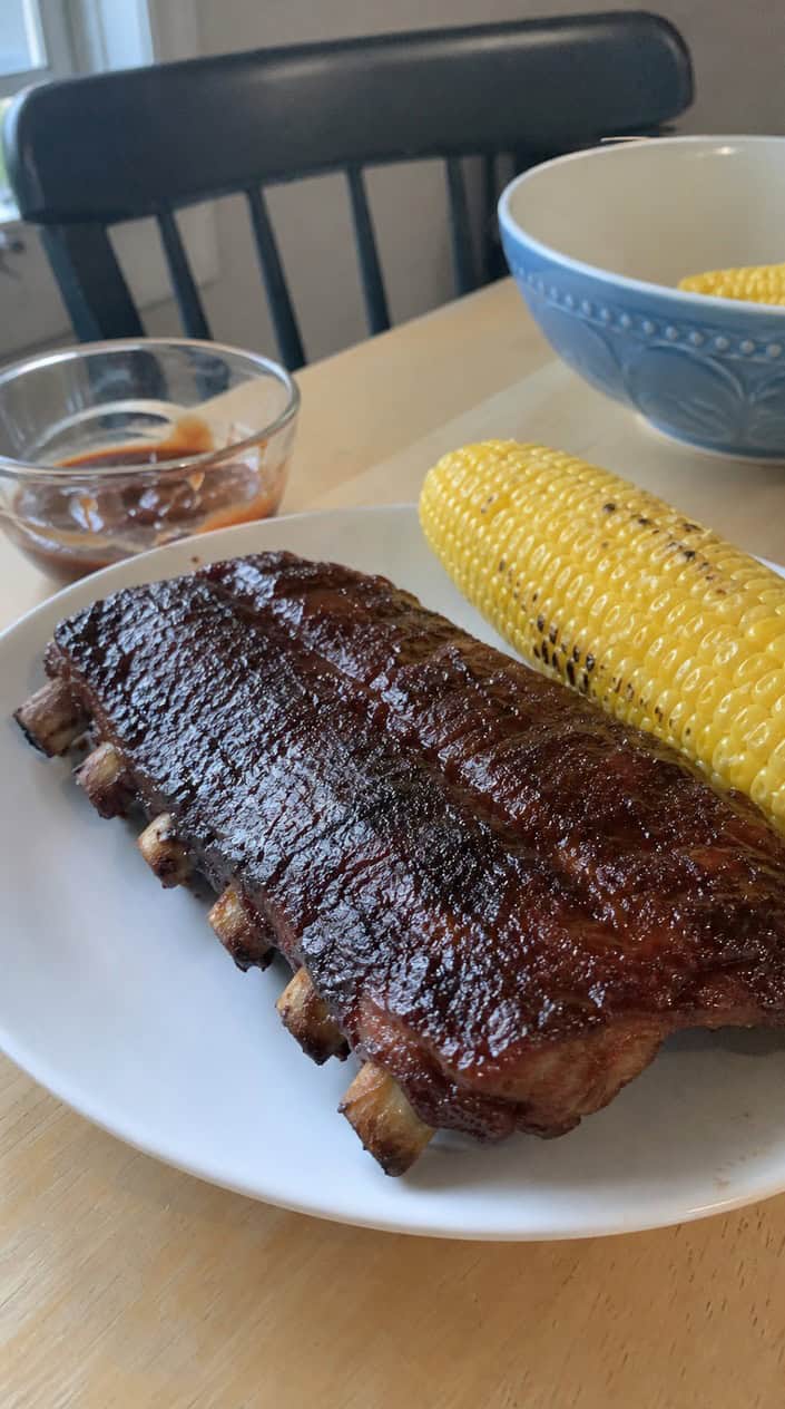 A platter of grilled BBQ baby back ribs with a smoky dry rub on a wooden table, surrounded by barbecue sauce and grilled corn, set in an outdoor backyard.