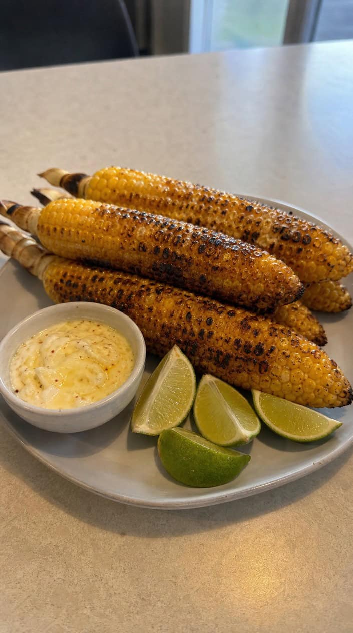 Grilled corn on the cob with lime-chili butter on a wooden board, accompanied by lime wedges and chili powder in a backyard setting.
