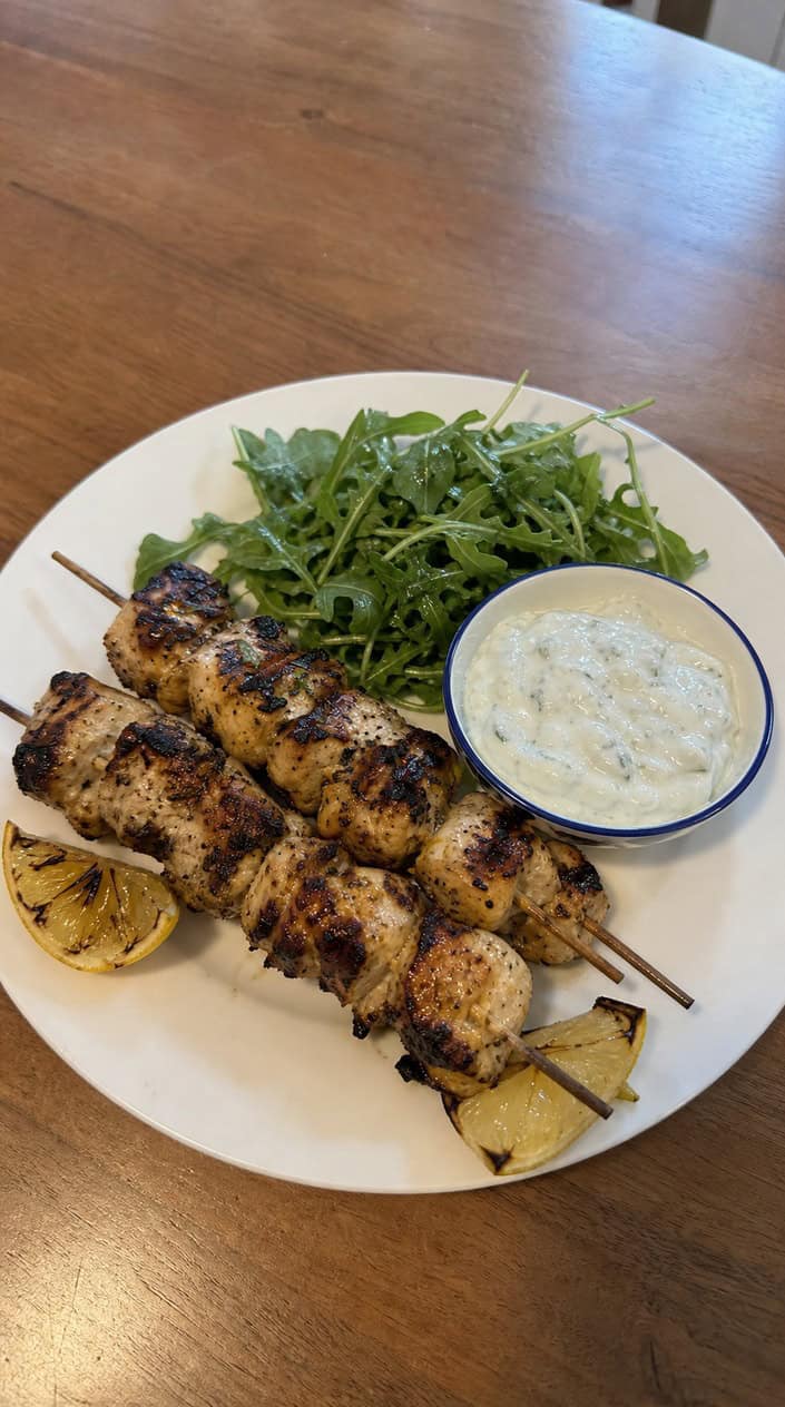Grilled lemon-garlic chicken skewers on a wooden table with a bowl of tzatziki sauce and lemon wedges.