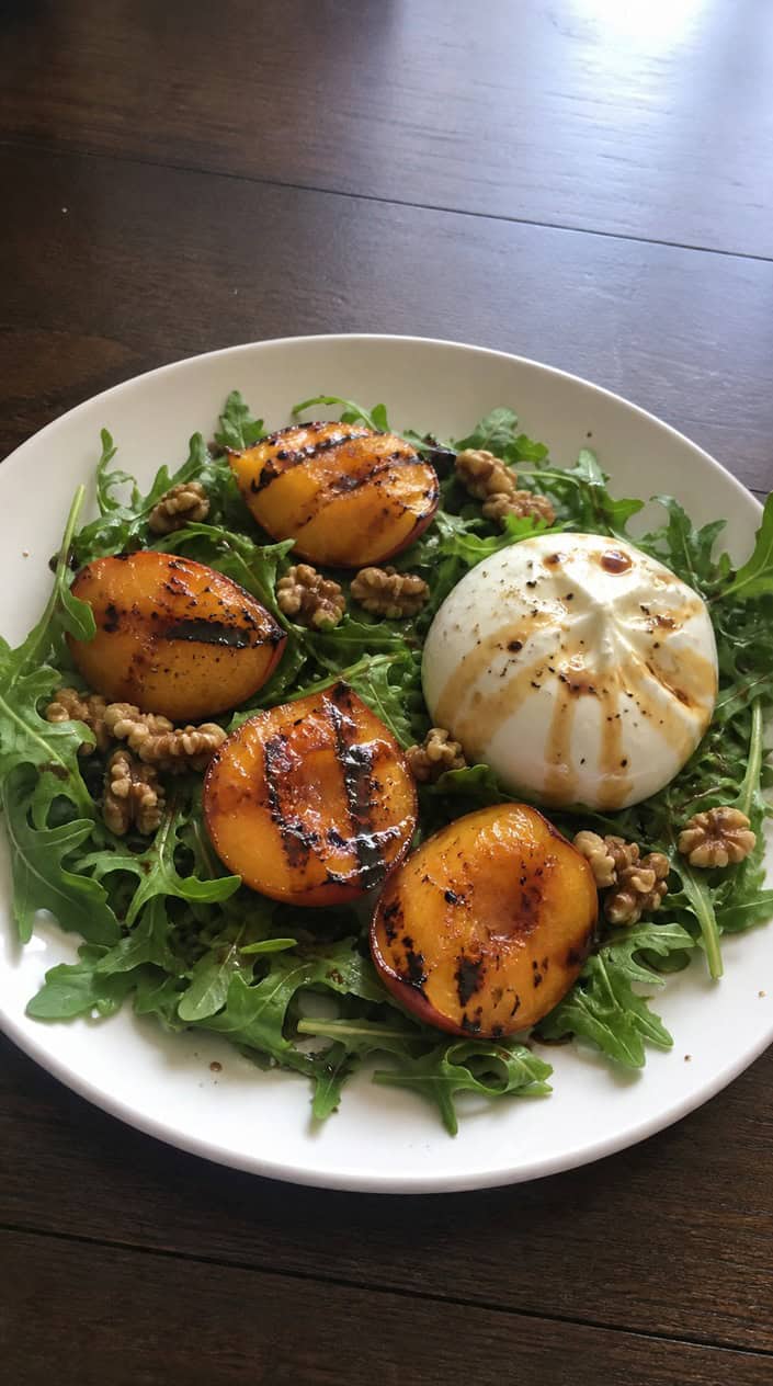 A plate of grilled peach and burrata salad with honey-balsamic dressing on a rustic table with summer sunlight.