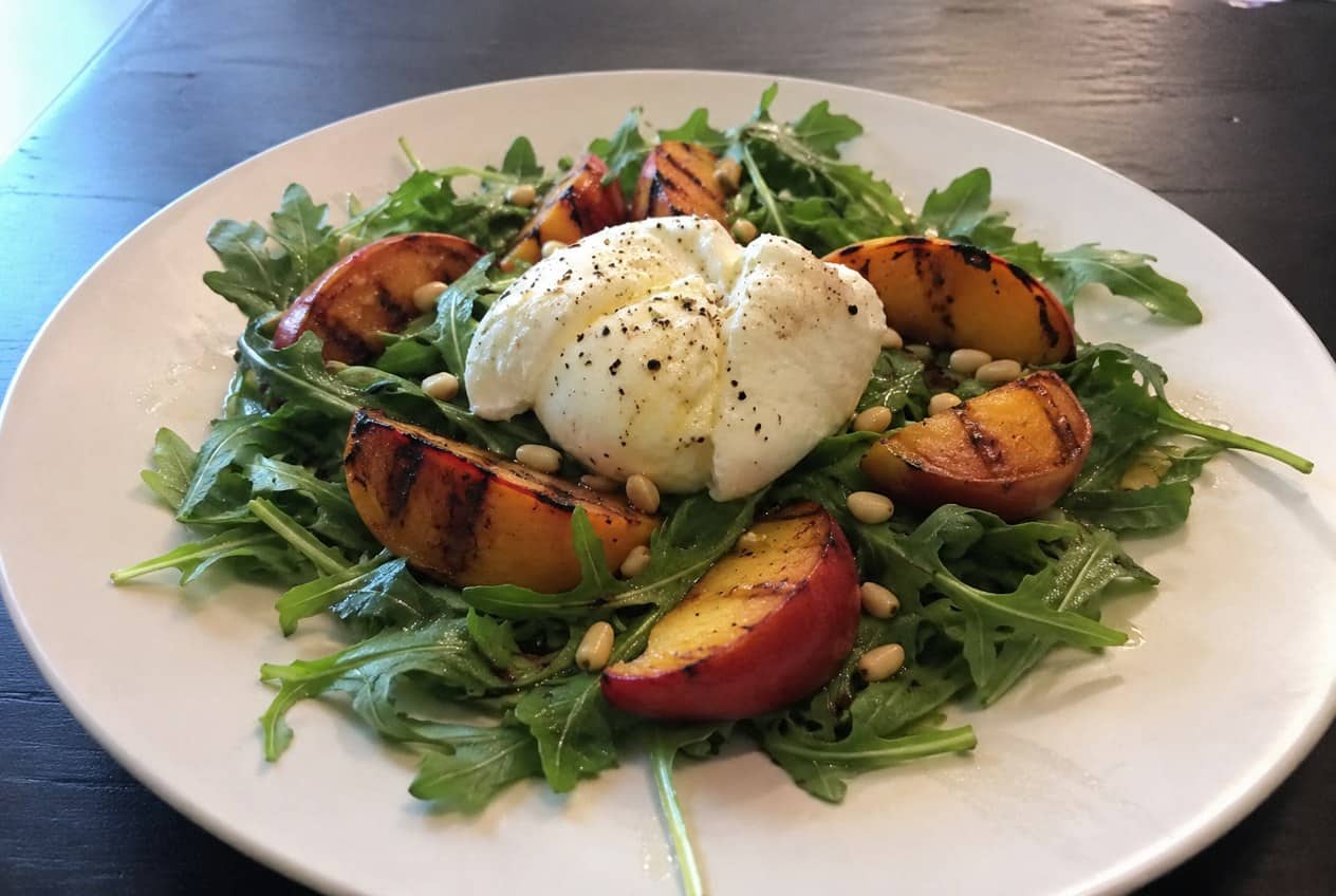 A white platter scattered with peppery arugula, caramelized grilled peach halves, torn burrata cheese, pine nuts, and a dark balsamic glaze drizzle.