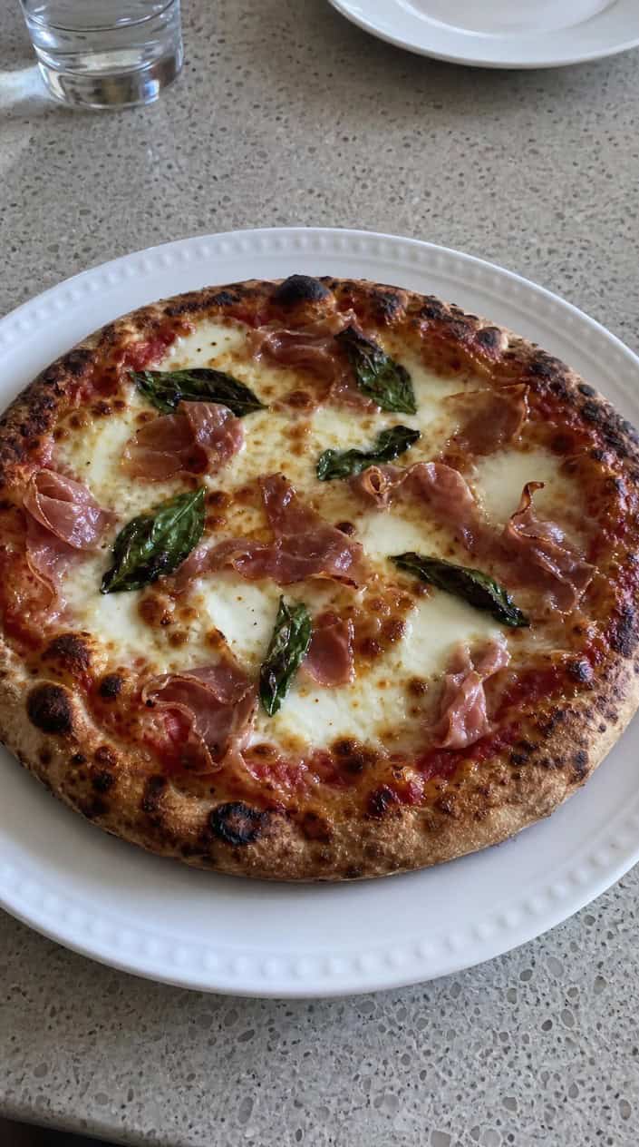 Grilled pizza topped with mozzarella, basil, and prosciutto cooking on a backyard grill outdoors.