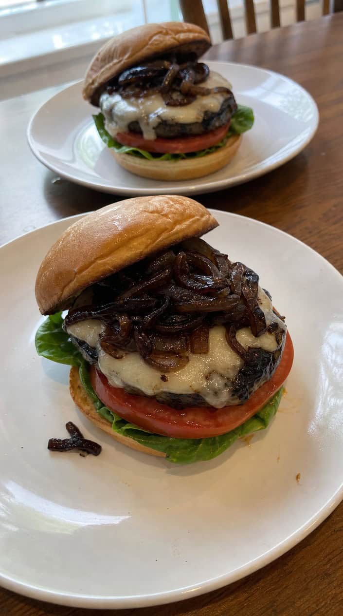 Two grilled portobello mushroom burgers with balsamic onions and fresh vegetables on a wooden table outdoors.