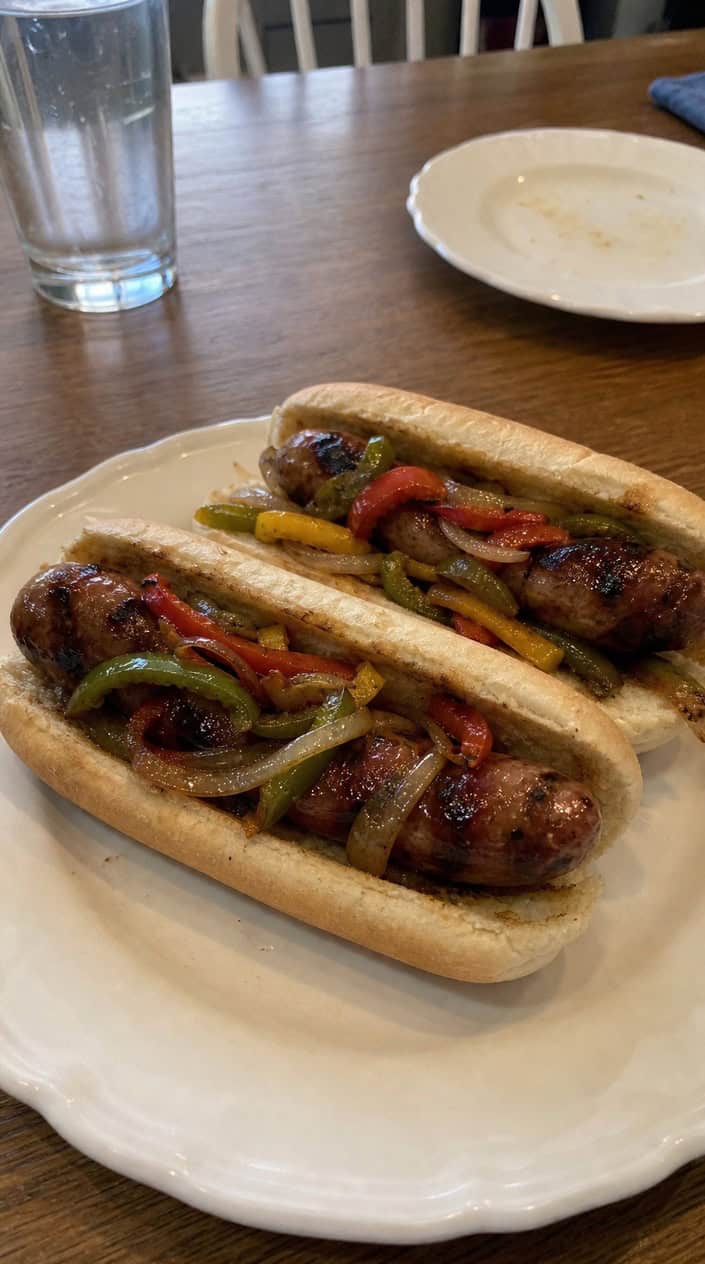 Grilled sausages and sautéed bell peppers served on hoagie rolls placed on a wooden picnic table in a backyard setting.