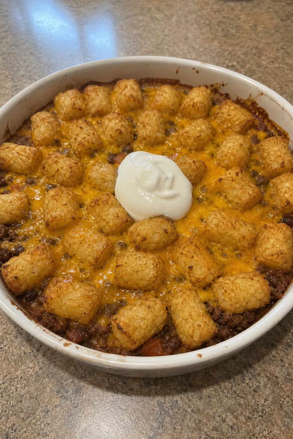 A baked casserole dish with a ground beef taco tater tot filling, the surface topped with melted orange cheddar and a scattering of green cilantro and white sour cream dollops.