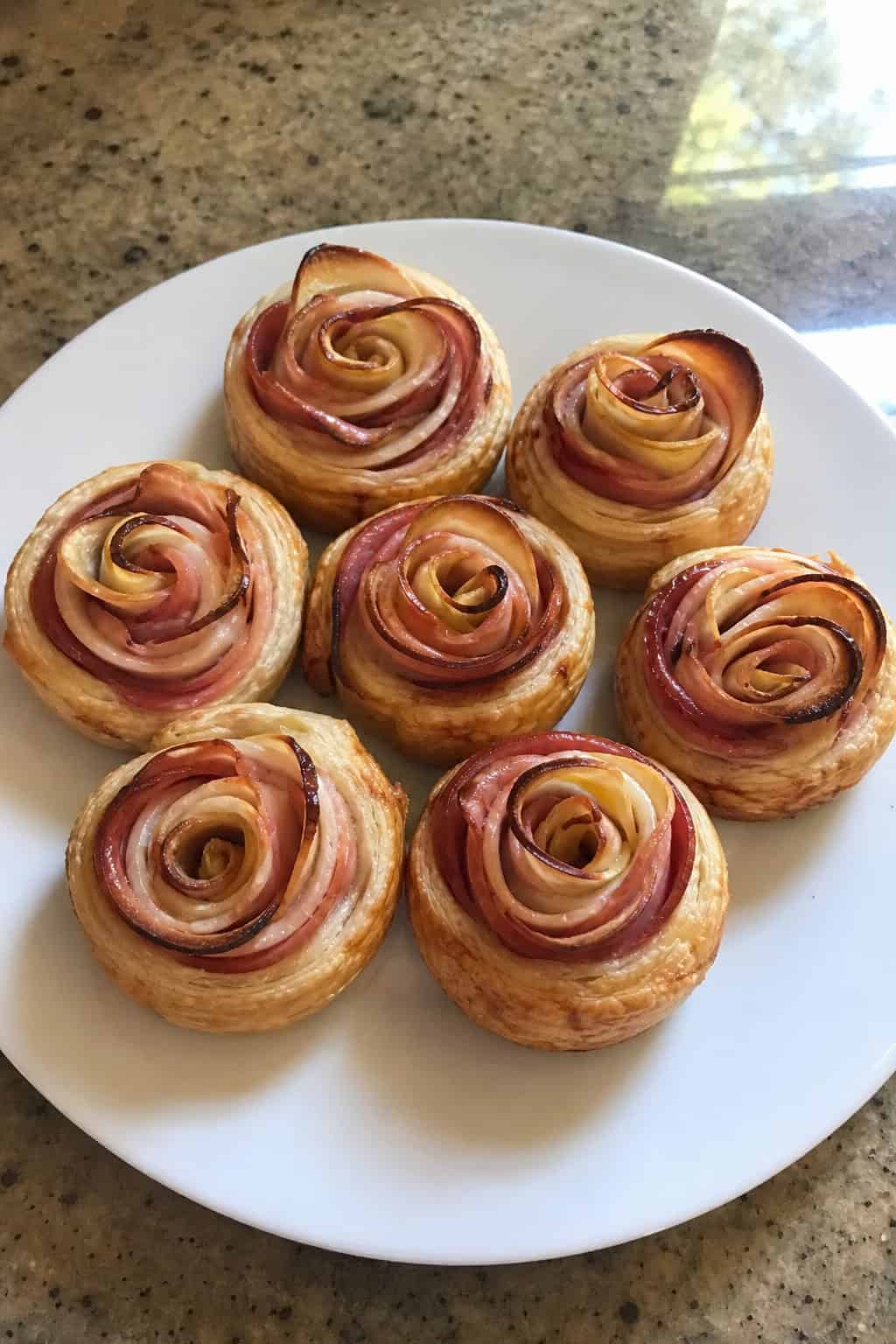 Plate of ham and apple puff pastry roses arranged on a wooden table with herbs and a small bowl of sauce in the background.