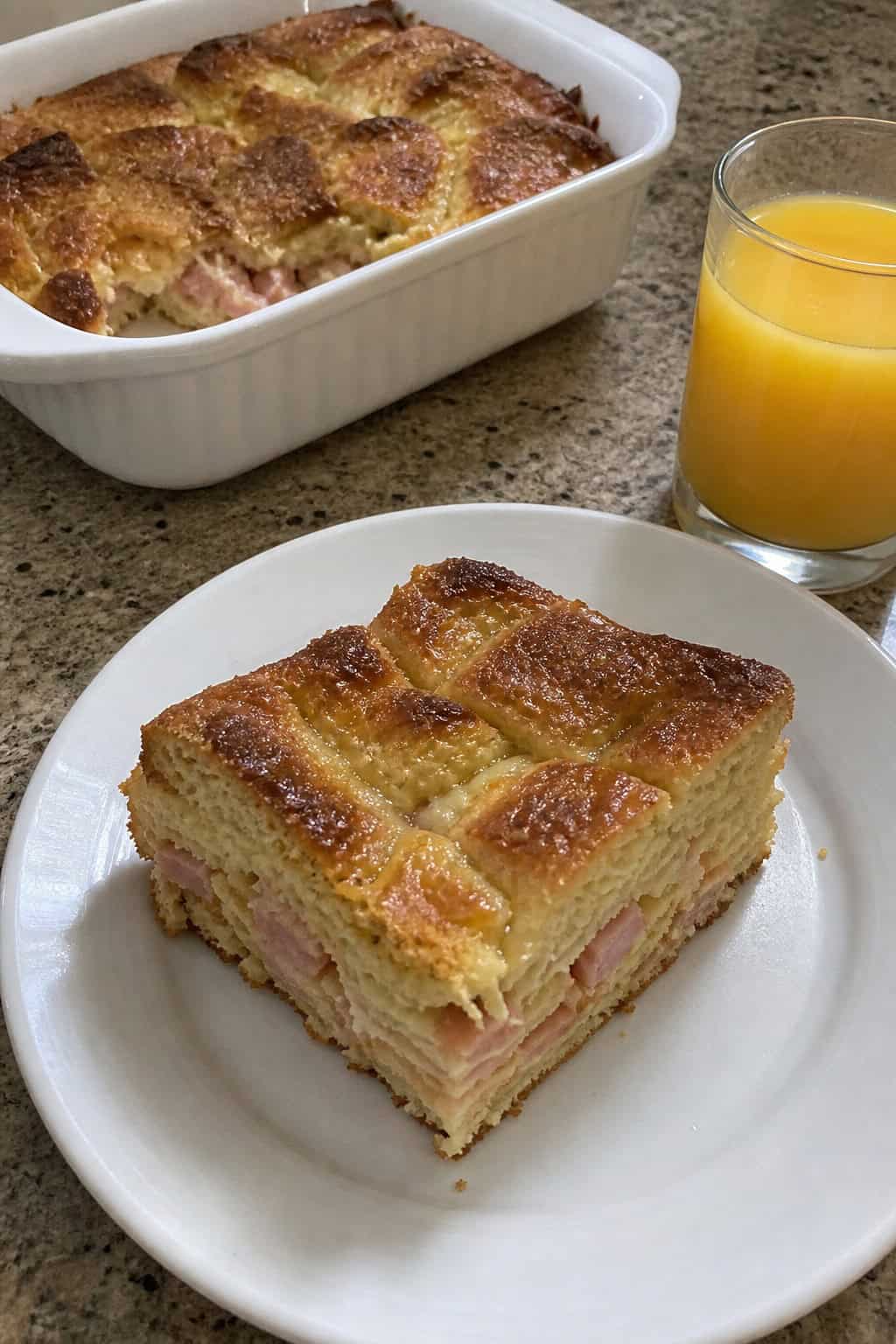 A plated slice of ham and Swiss French toast strata on a white plate garnished with fresh herbs, with a glass of orange juice and a small bowl of berries visible on the wooden table beside it.