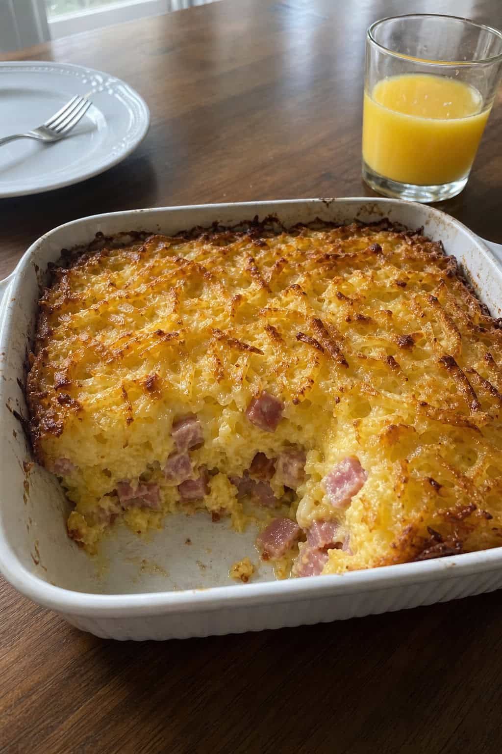 A ham and cheese hashbrown casserole in a round ceramic dish with a golden, slightly crisped top, sitting on a wood table with a glass of orange juice and a fork resting nearby.