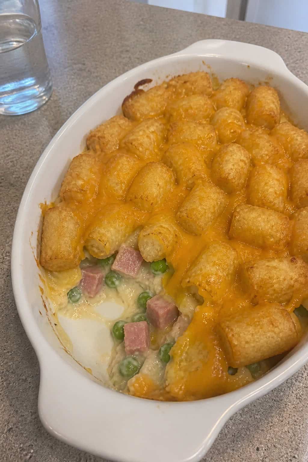 A baked tater tot casserole with pink-edged diced ham, bright green peas, and melted cheddar visible at the casserole edges, with golden tots covering the top in a ceramic dish on a wooden table.