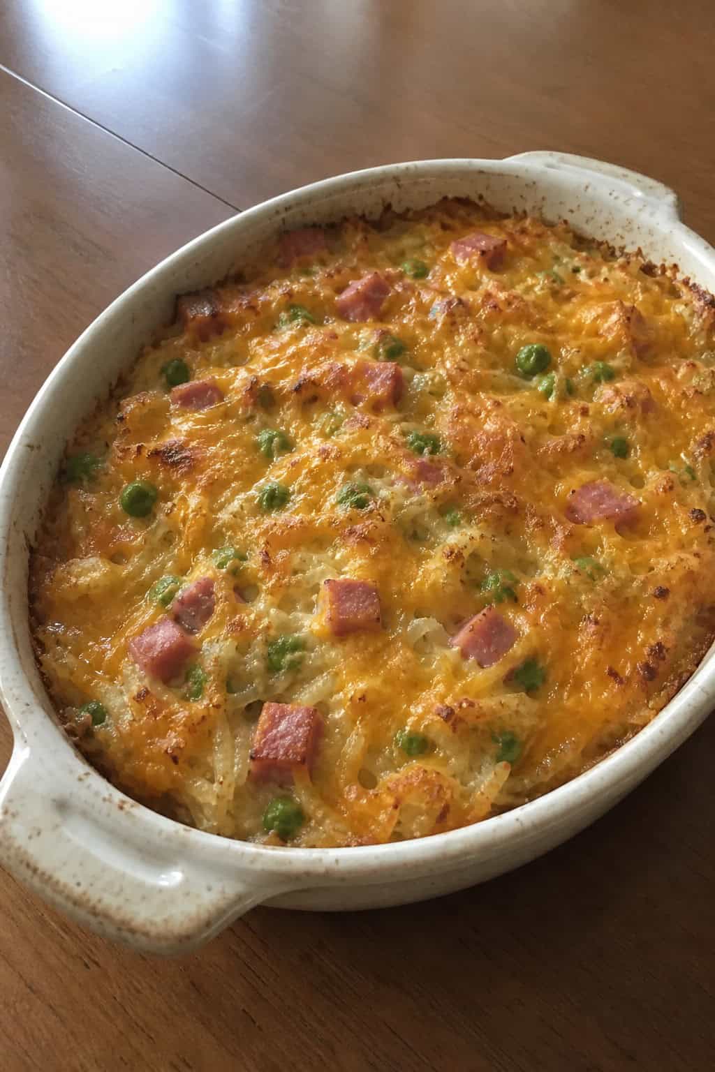 A baked ham, pea, and cheddar hashbrown casserole in a ceramic dish on a wooden table with a serving spatula and a bowl of peas nearby.