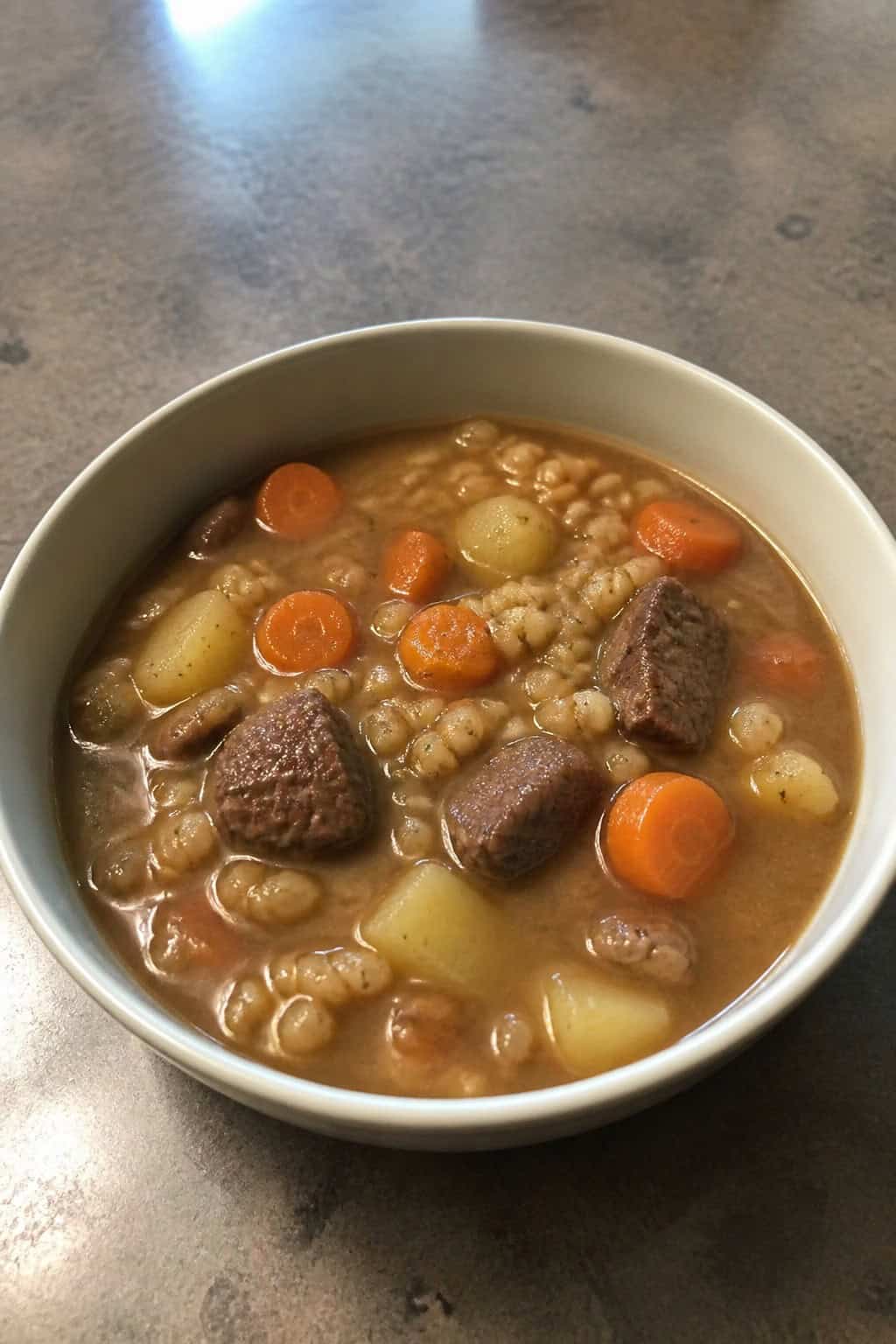 A bowl of beef barley and root vegetable soup on a wooden table with fresh vegetables and herbs nearby.