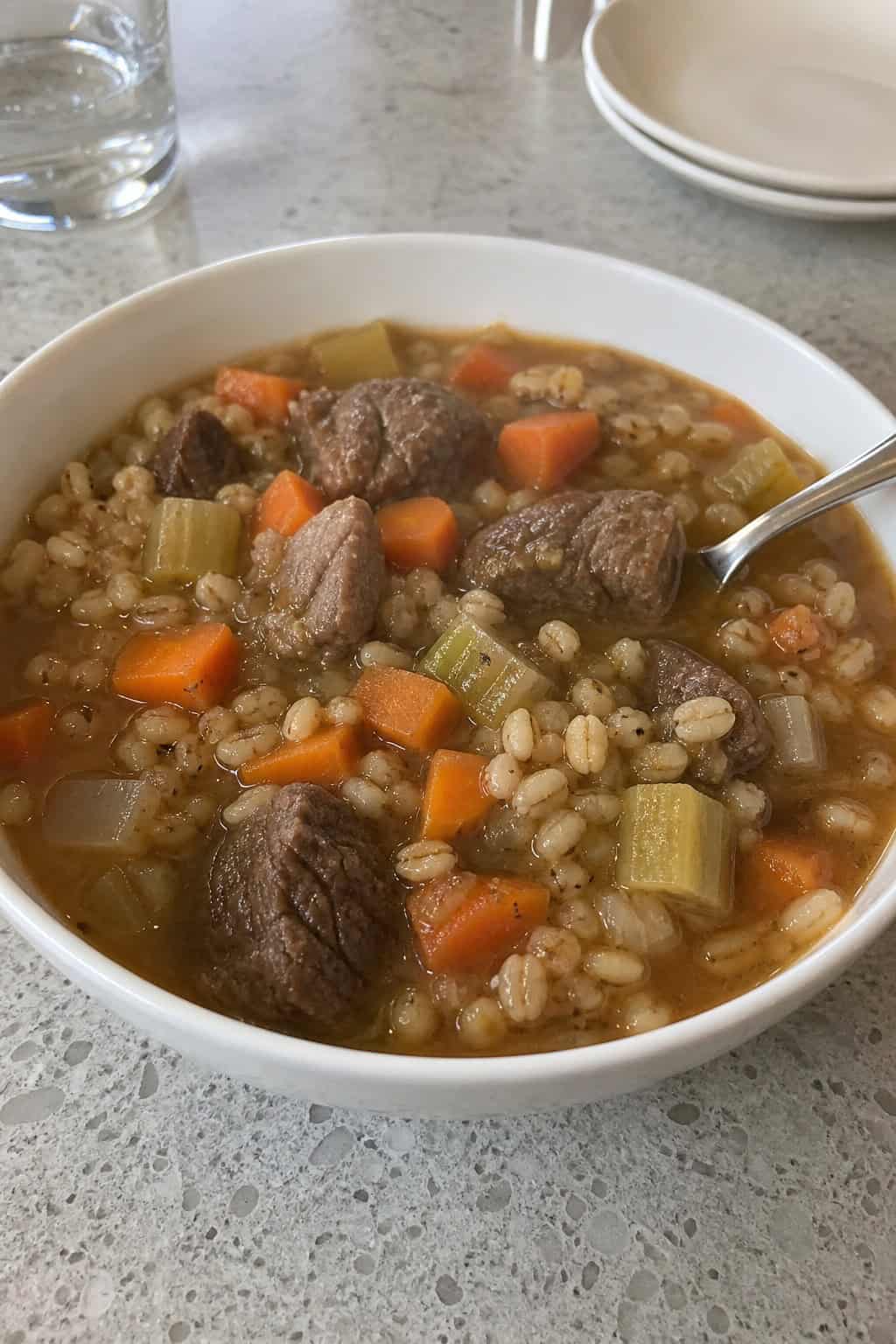 A bowl of steaming beef barley soup with diced vegetables on a wooden table, surrounded by fresh ingredients and a wooden spoon.