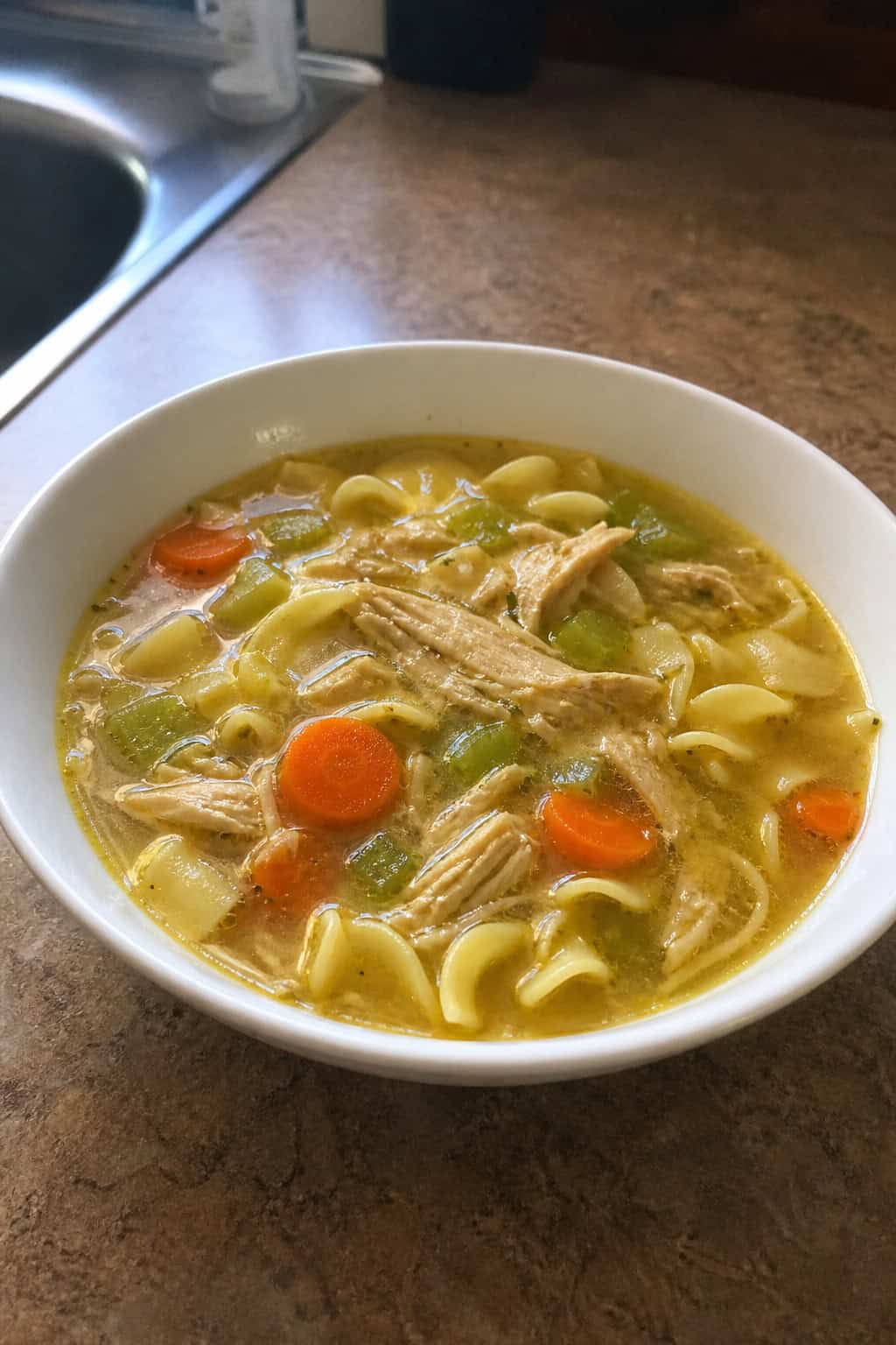 A crockpot filled with steaming chicken noodle soup on a wooden table surrounded by fresh vegetables and herbs.