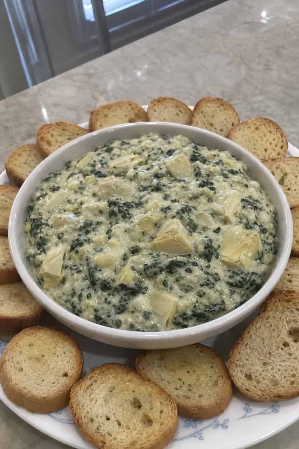 A bowl of spinach artichoke dip with toasted crostini arranged around it on a wooden board.