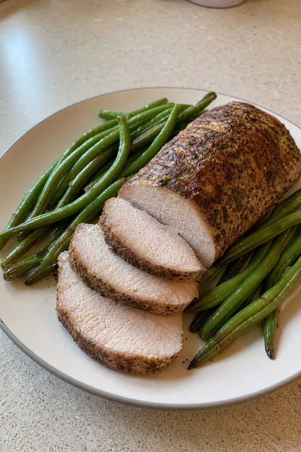 A sliced herb-crusted pork tenderloin surrounded by tender roasted green beans and thin lemon rounds on a wooden cutting board set on a kitchen countertop.