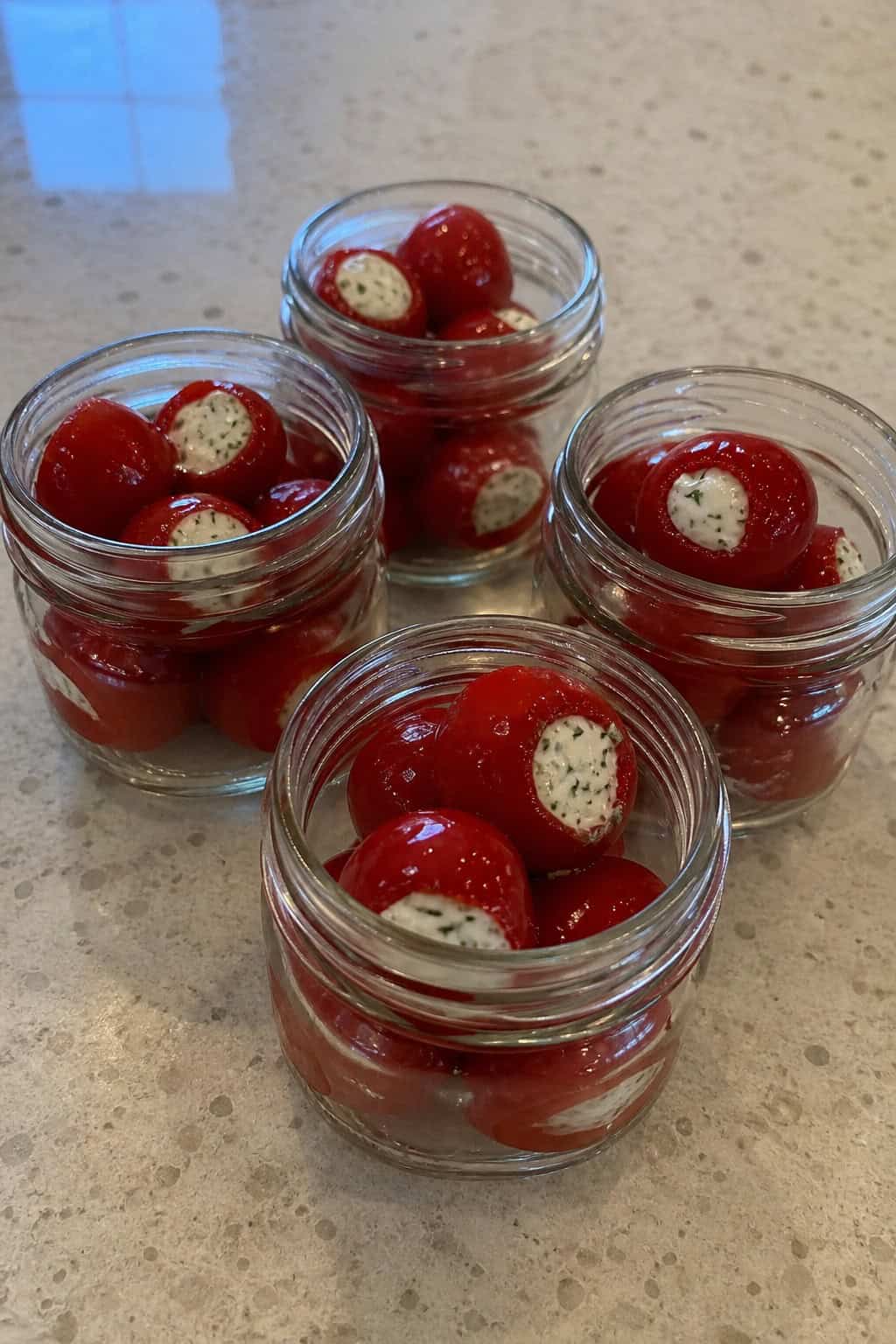 Close-up of glass jars filled with red peppadew peppers stuffed with herbed goat cheese, arranged on a wooden surface with fresh herbs around them.