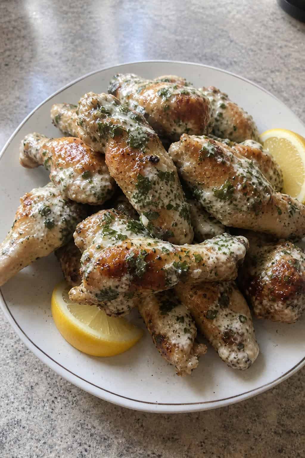A plate of crispy chicken wings coated in herbed lemon-garlic yogurt sauce with lemon slices and fresh herbs on a wooden table.
