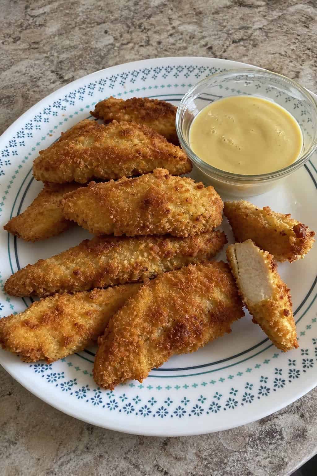 Crispy golden chicken tenders on a white plate next to a small bowl of dipping sauce and a few fresh vegetable pieces.