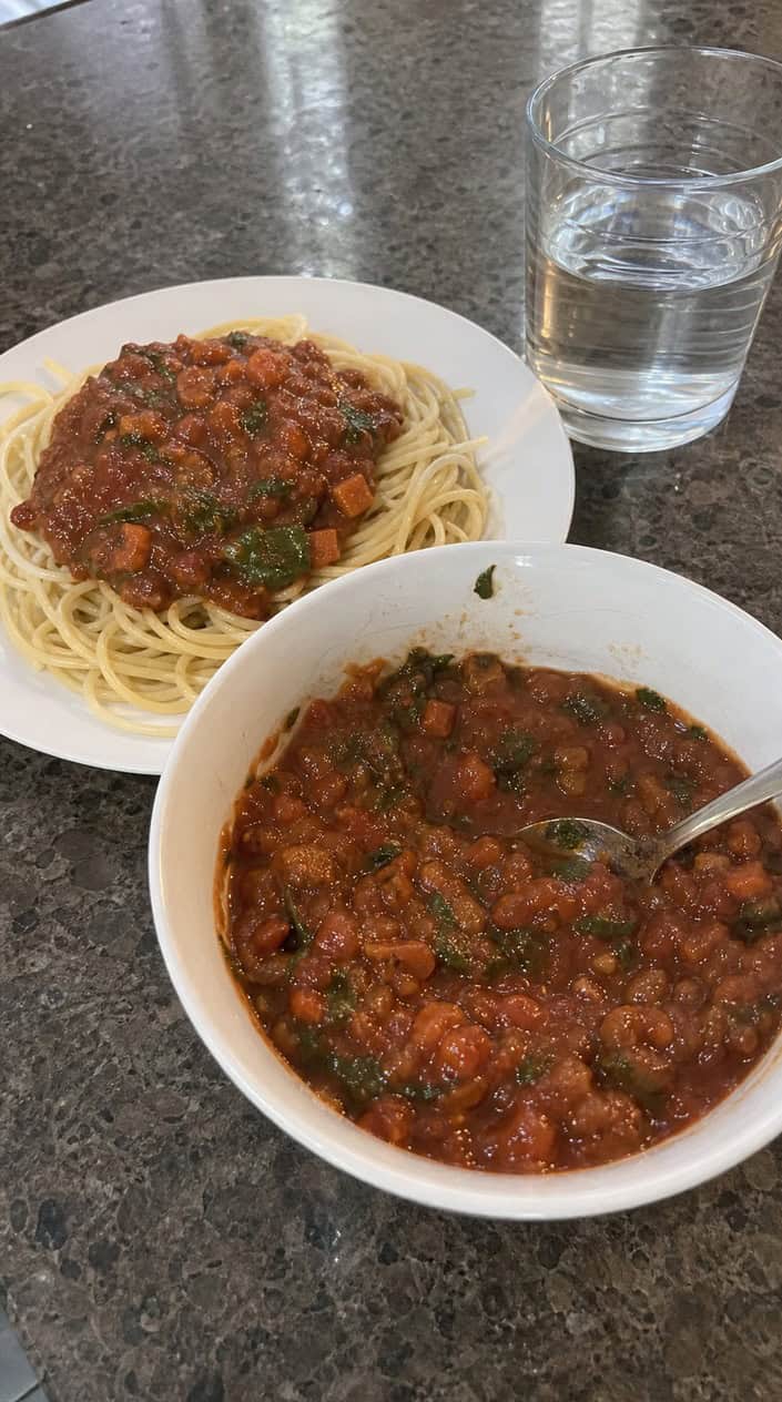 A bowl of homemade spaghetti sauce with hidden vegetables on a wooden countertop surrounded by fresh ingredients and a plate of spaghetti pasta.