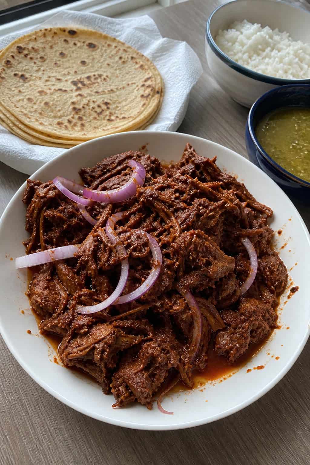 Plate of shredded guajillo steak carnitas garnished with cilantro and onions, with tortillas, salsa, and lime wedges on a wooden table.