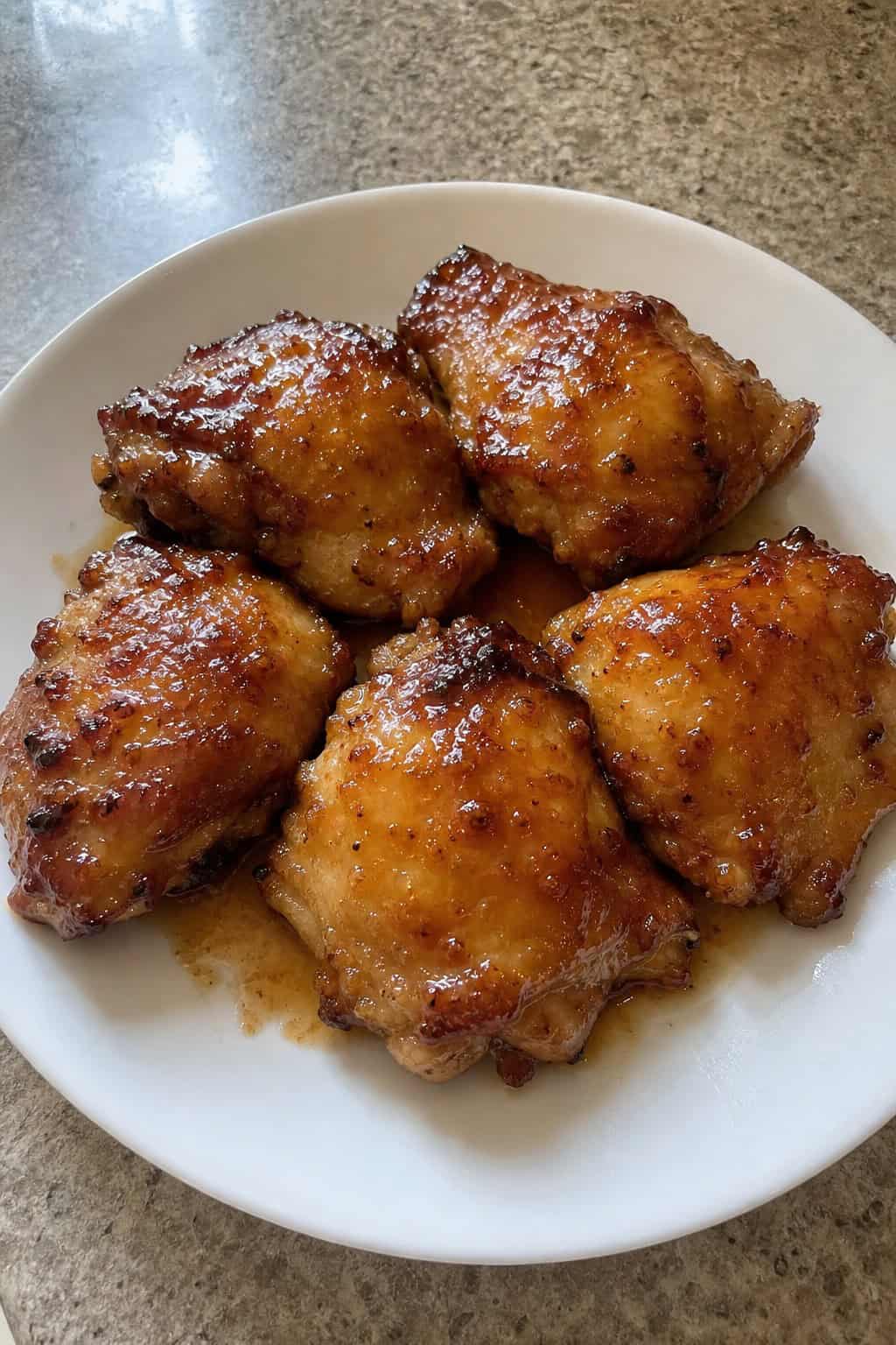 Close-up of honey garlic chicken thighs with crispy skin on a plate, garnished with herbs, with small bowls of ingredients nearby on a wooden table.