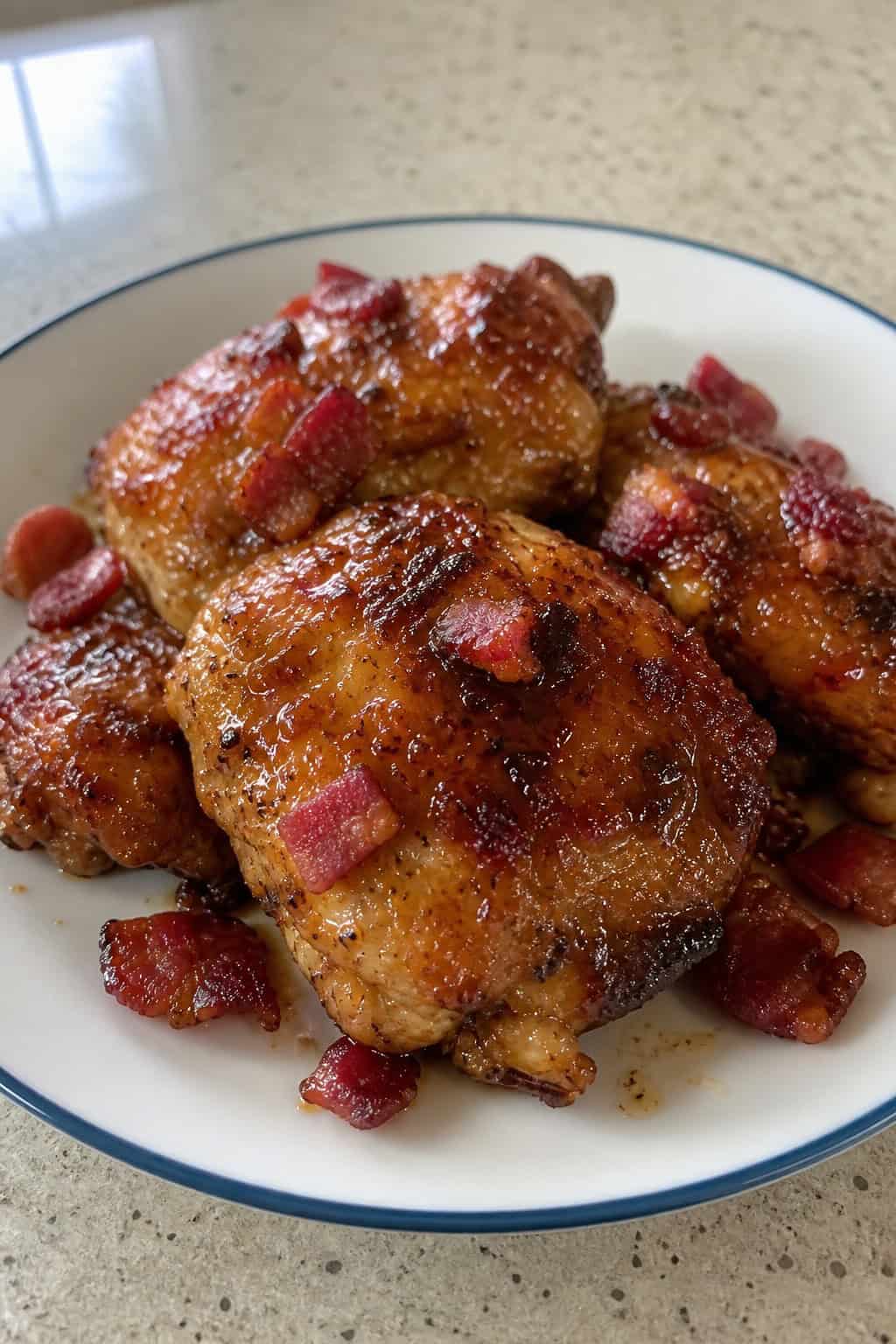 A plate of honey garlic chicken thighs topped with crispy bacon and maple glaze, garnished with fresh herbs on a wooden table.