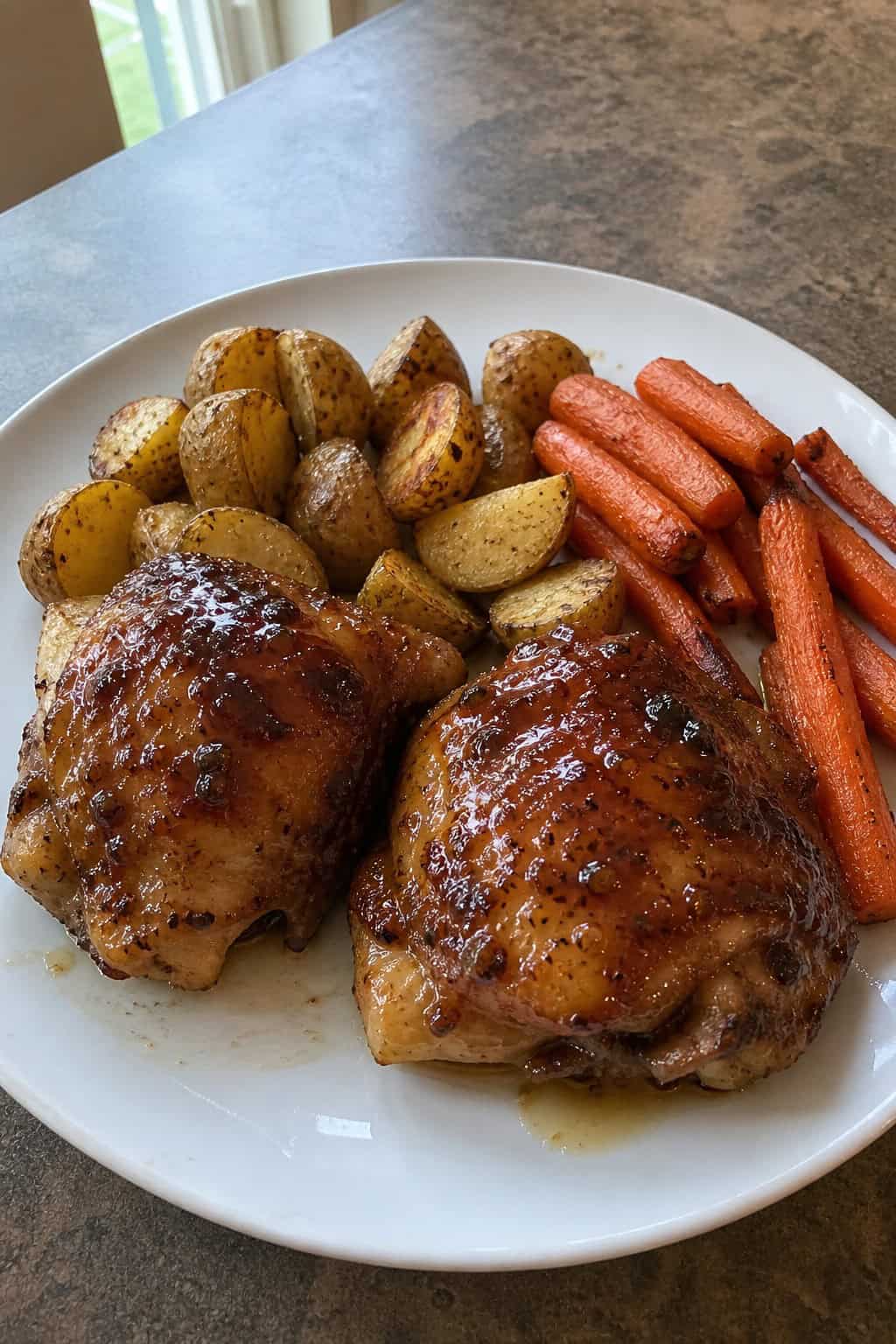 A plate of honey garlic chicken thighs with roasted carrots and potatoes on a wooden table.