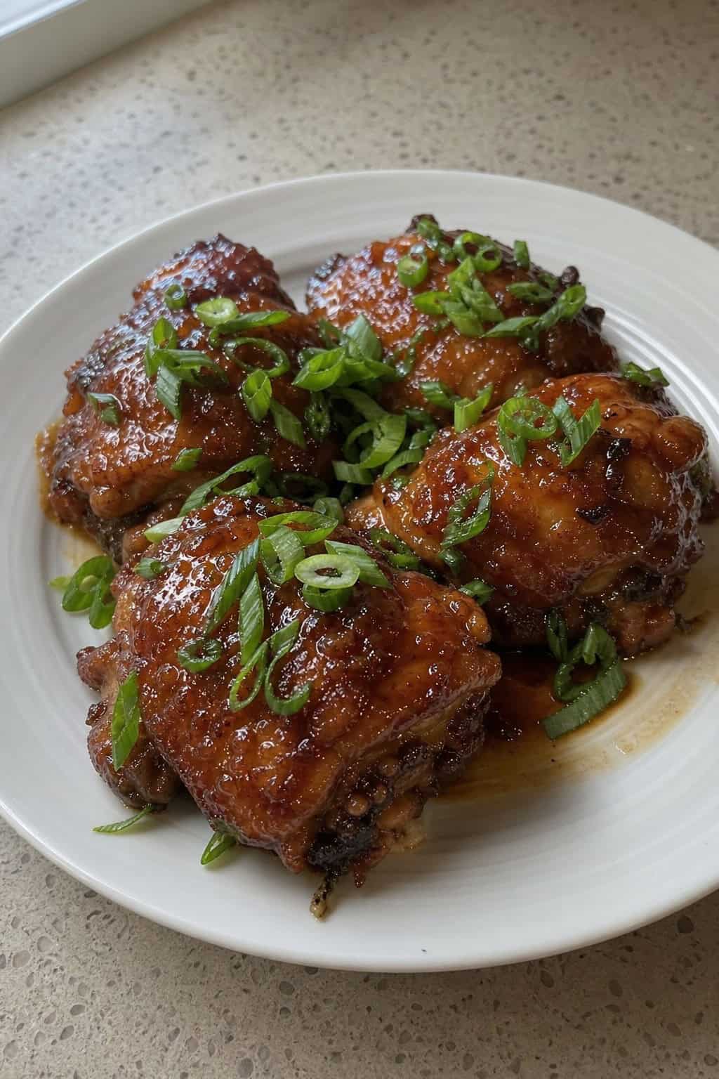 A plate of honey garlic chicken thighs glazed with hoisin sauce and topped with sliced scallions on a wooden table.