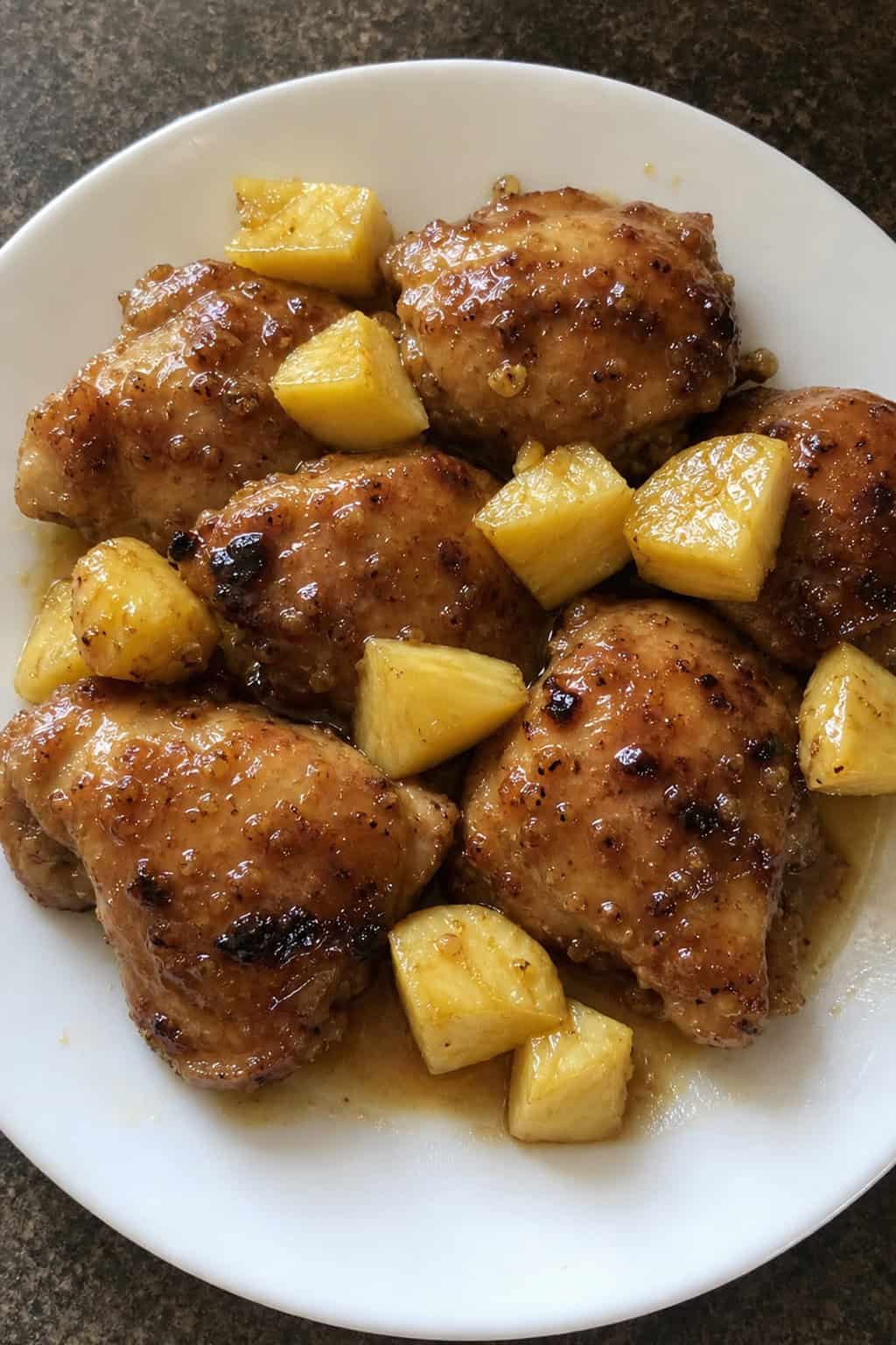 A plate of honey garlic chicken thighs garnished with pineapple chunks and herbs on a wooden table.