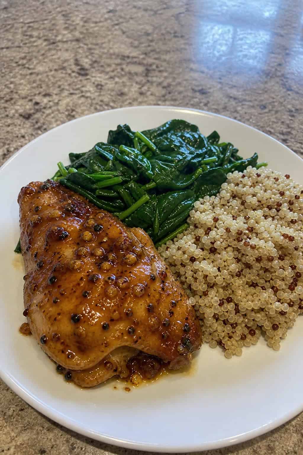 A plate with honey garlic chicken thighs, quinoa, and sautéed spinach on a wooden table.