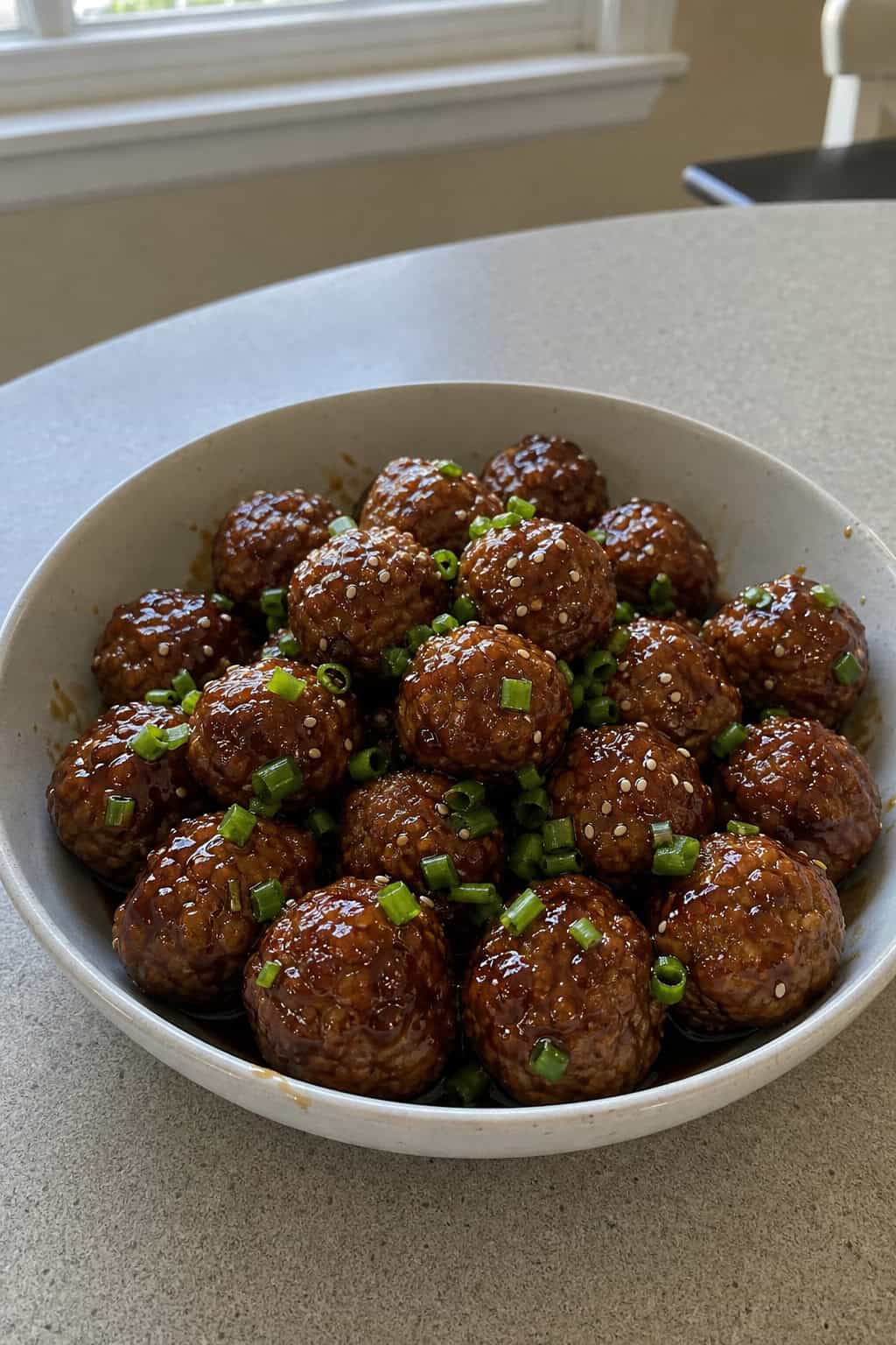 A bowl of honey-garlic cocktail meatballs garnished with green onions and sesame seeds on a wooden table.