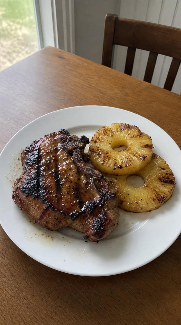 Honey-garlic pork chops with grilled pineapple on a plate set on a wooden table outdoors with a charcoal grill and greenery in the background.