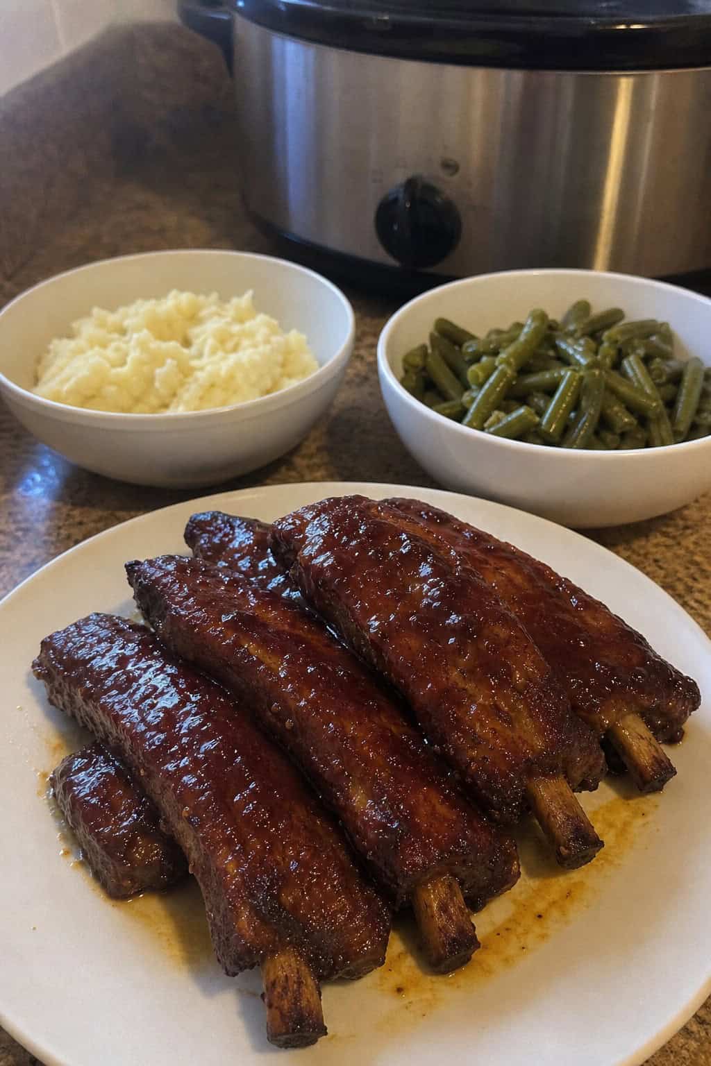 Plate of honey garlic ribs with side dishes on a wooden table and a slow cooker in the background.